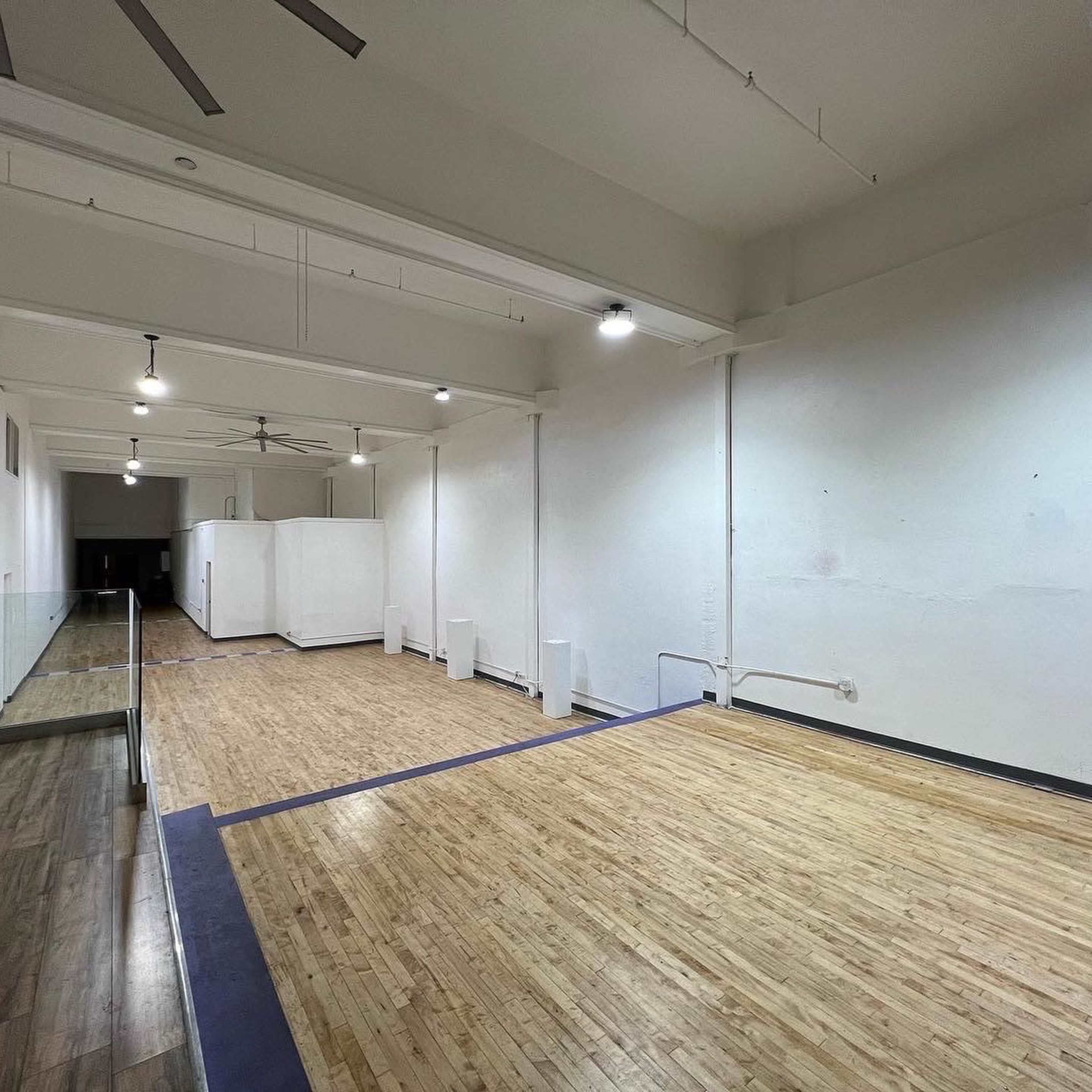 A spacious, empty room with wooden flooring and white walls, featuring ceiling fans and minimal furnishings.