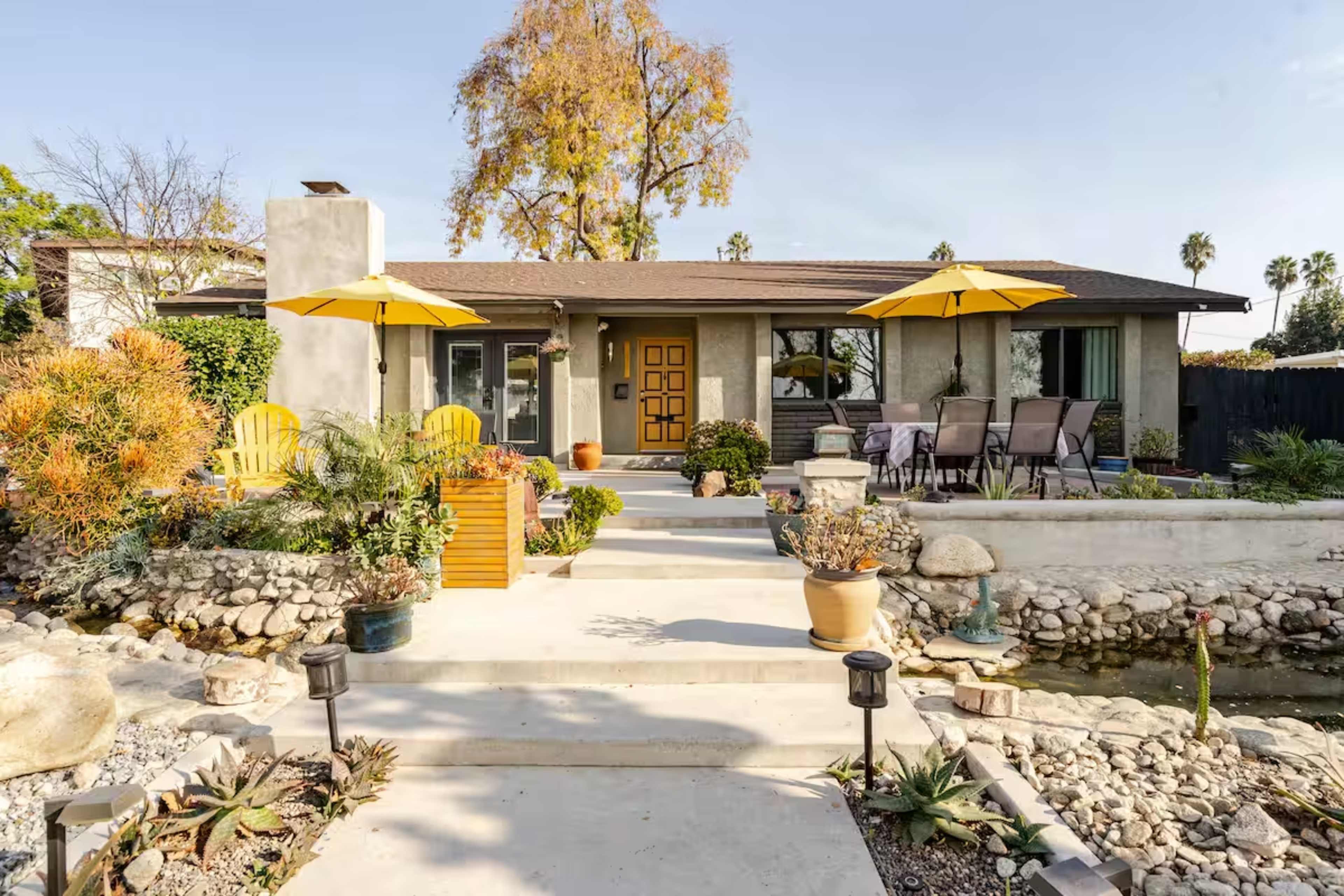 A single-story house features a landscaped front yard with stone pathways, outdoor seating, and yellow umbrellas.