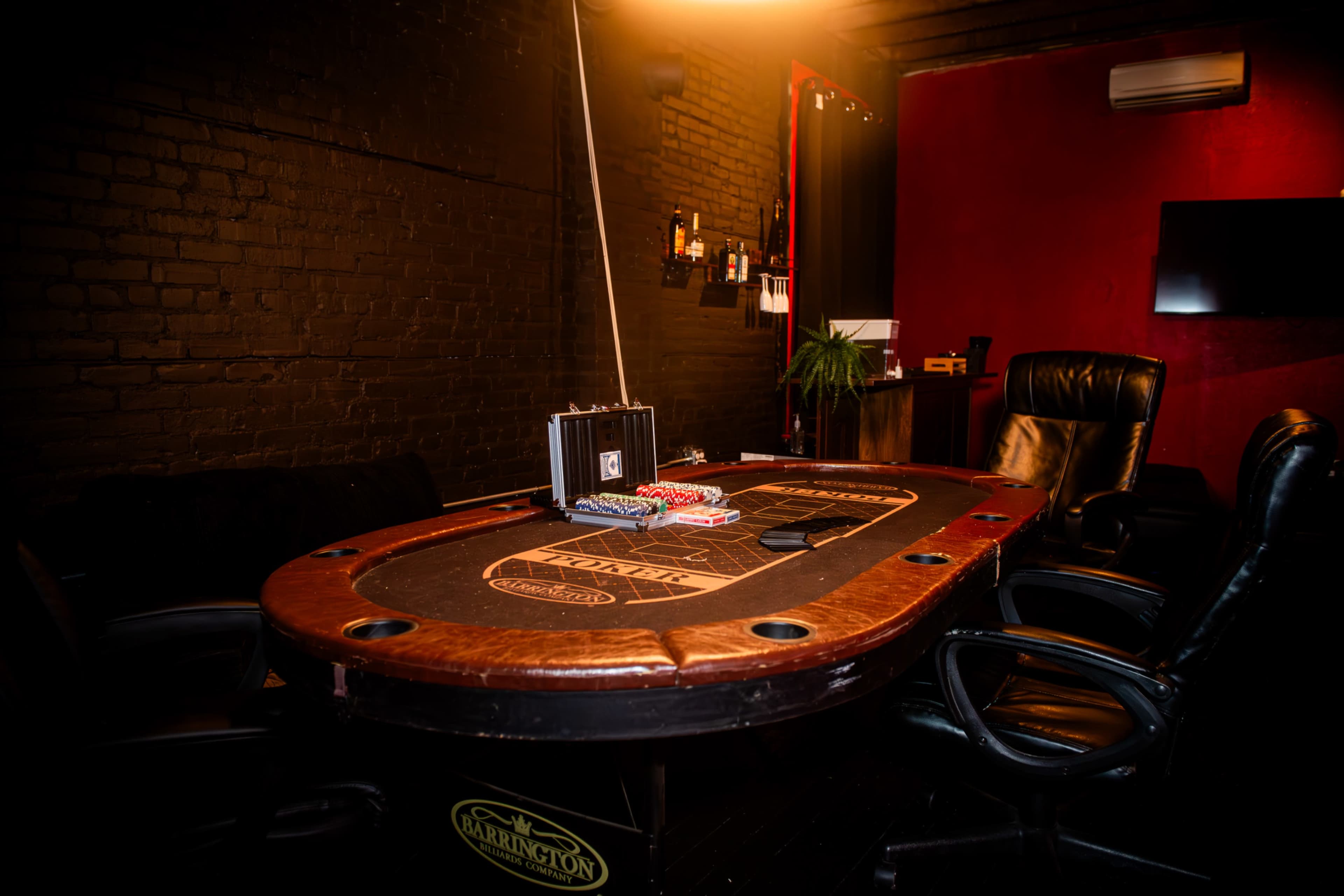 The image shows a poker table with playing cards and chips set in a room featuring dark walls and leather chairs.