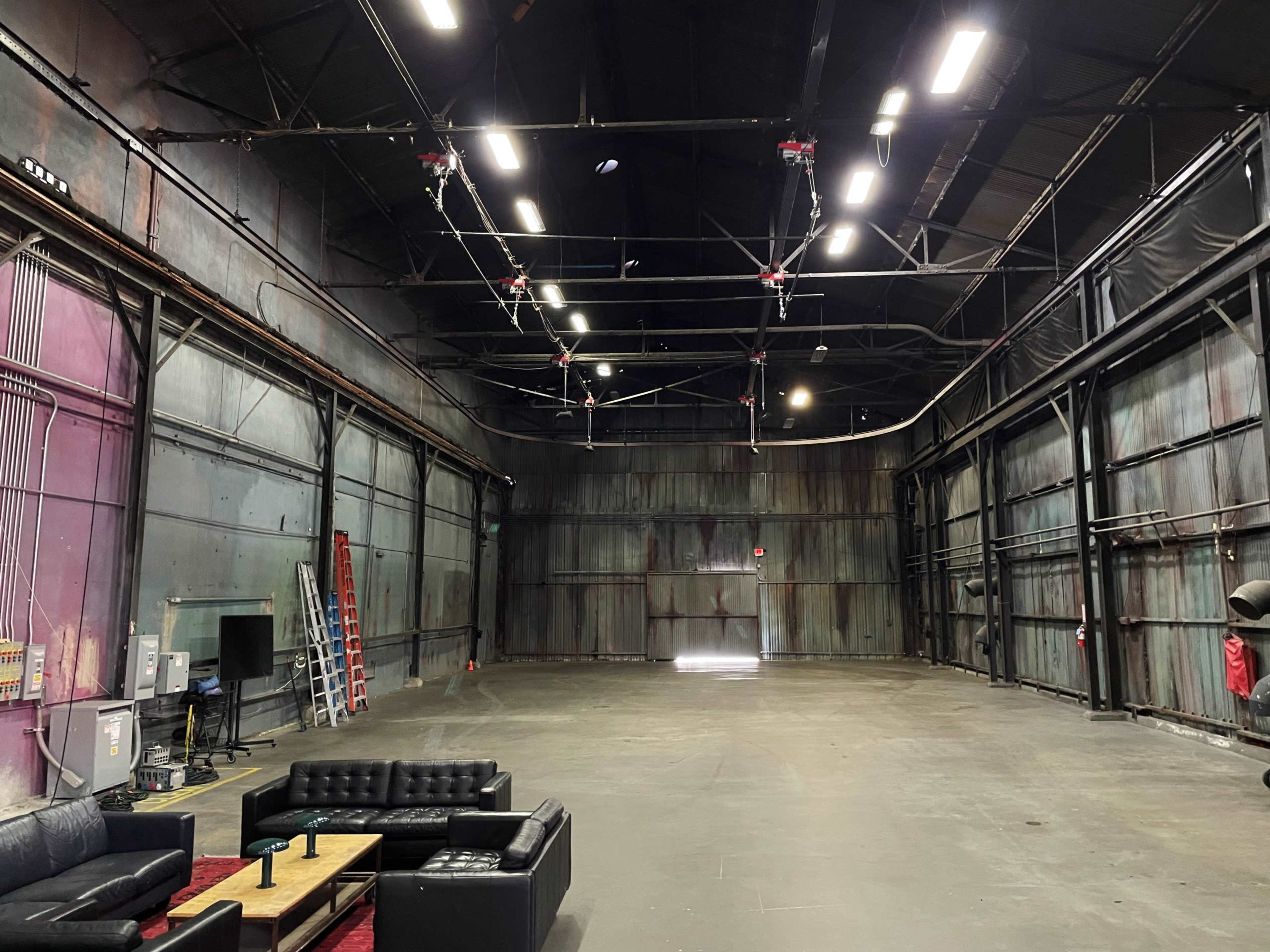 The image shows a large, empty industrial space with metal walls, overhead equipment, a ladder, and a seating area featuring a black sofa and a coffee table.