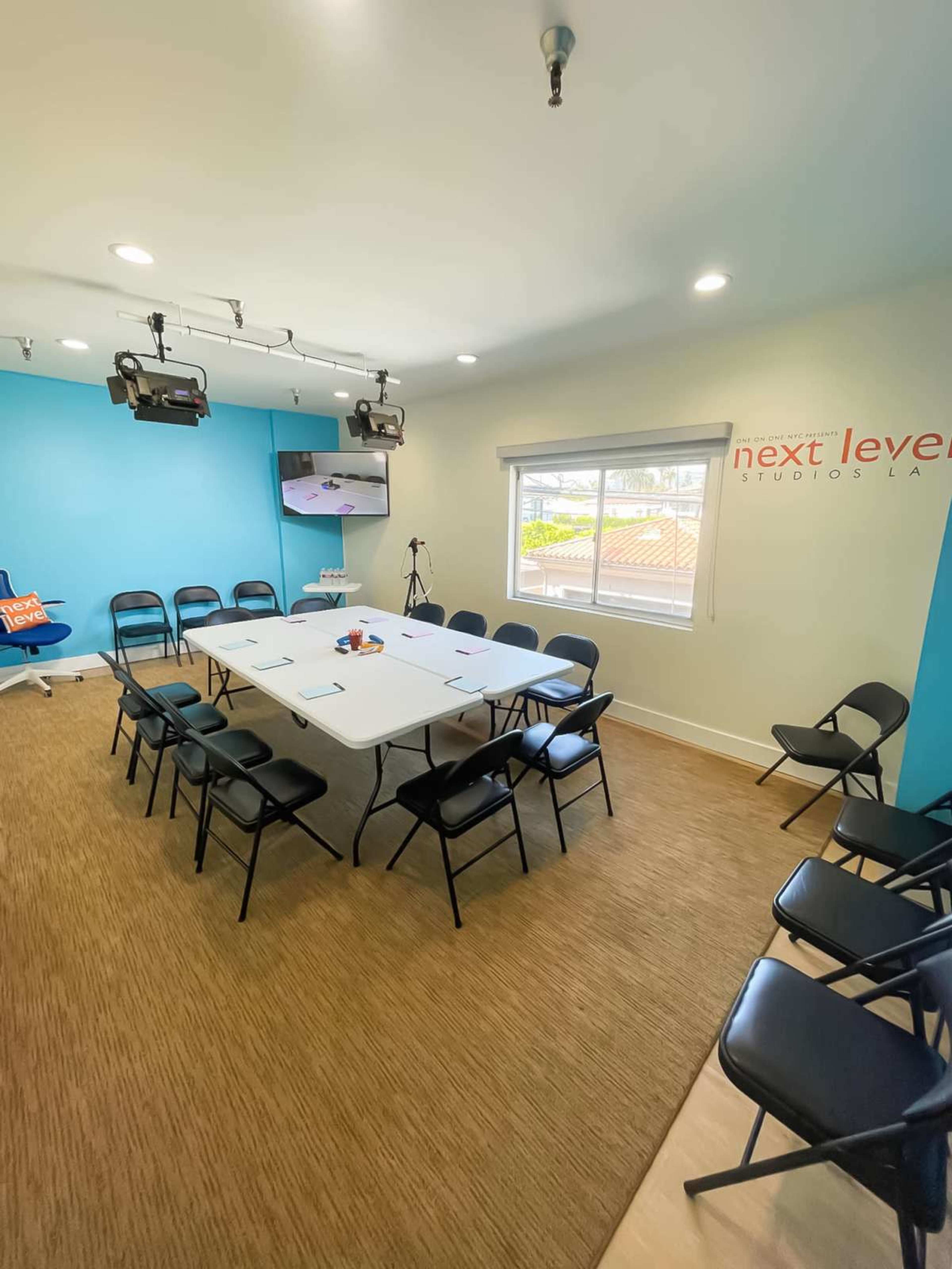 The image shows a brightly lit conference room with a large table surrounded by black chairs, featuring a television mounted on the wall and a blue accent wall.