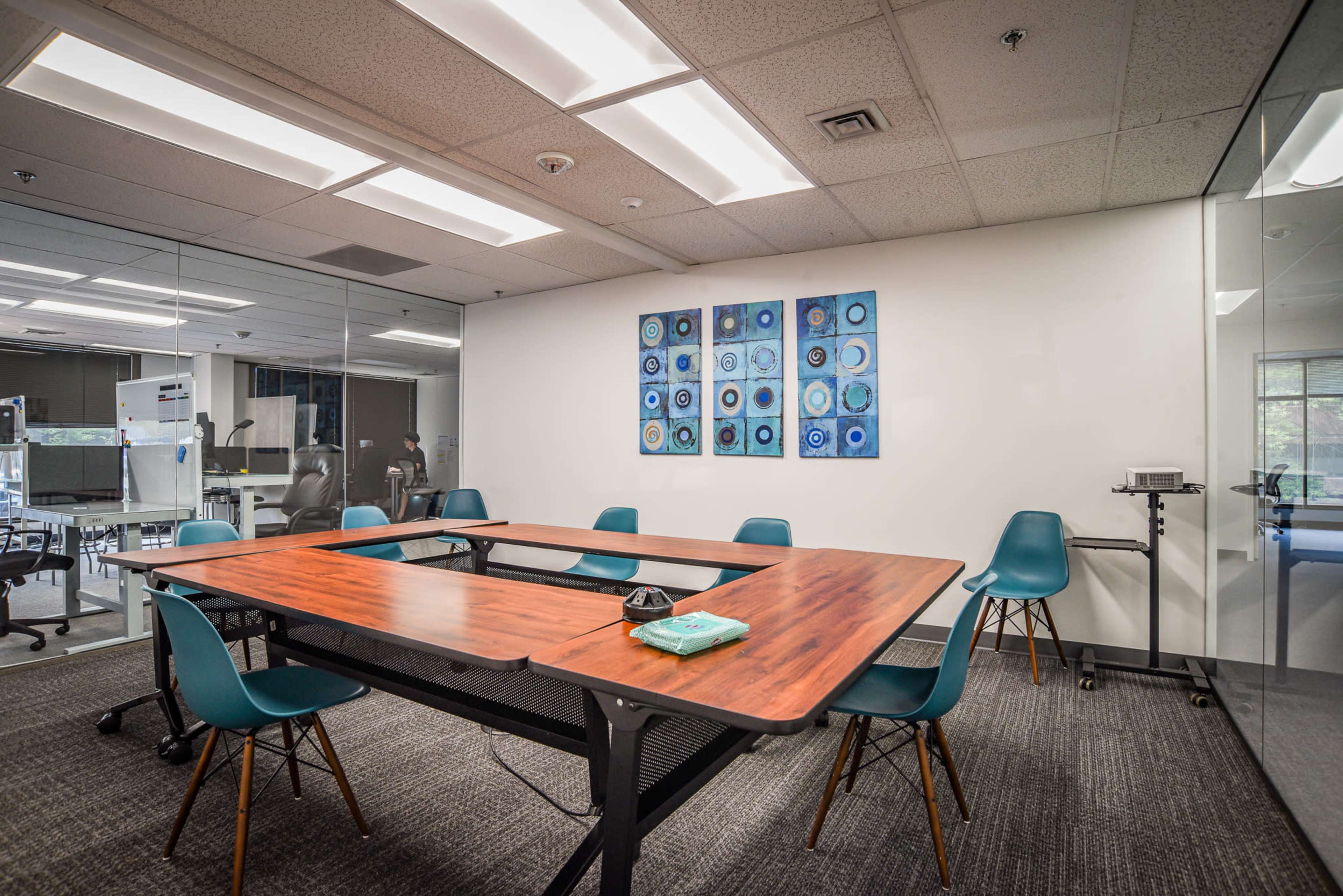 A modern conference room with a wooden table surrounded by teal chairs, and a large wall with framed blue artwork.