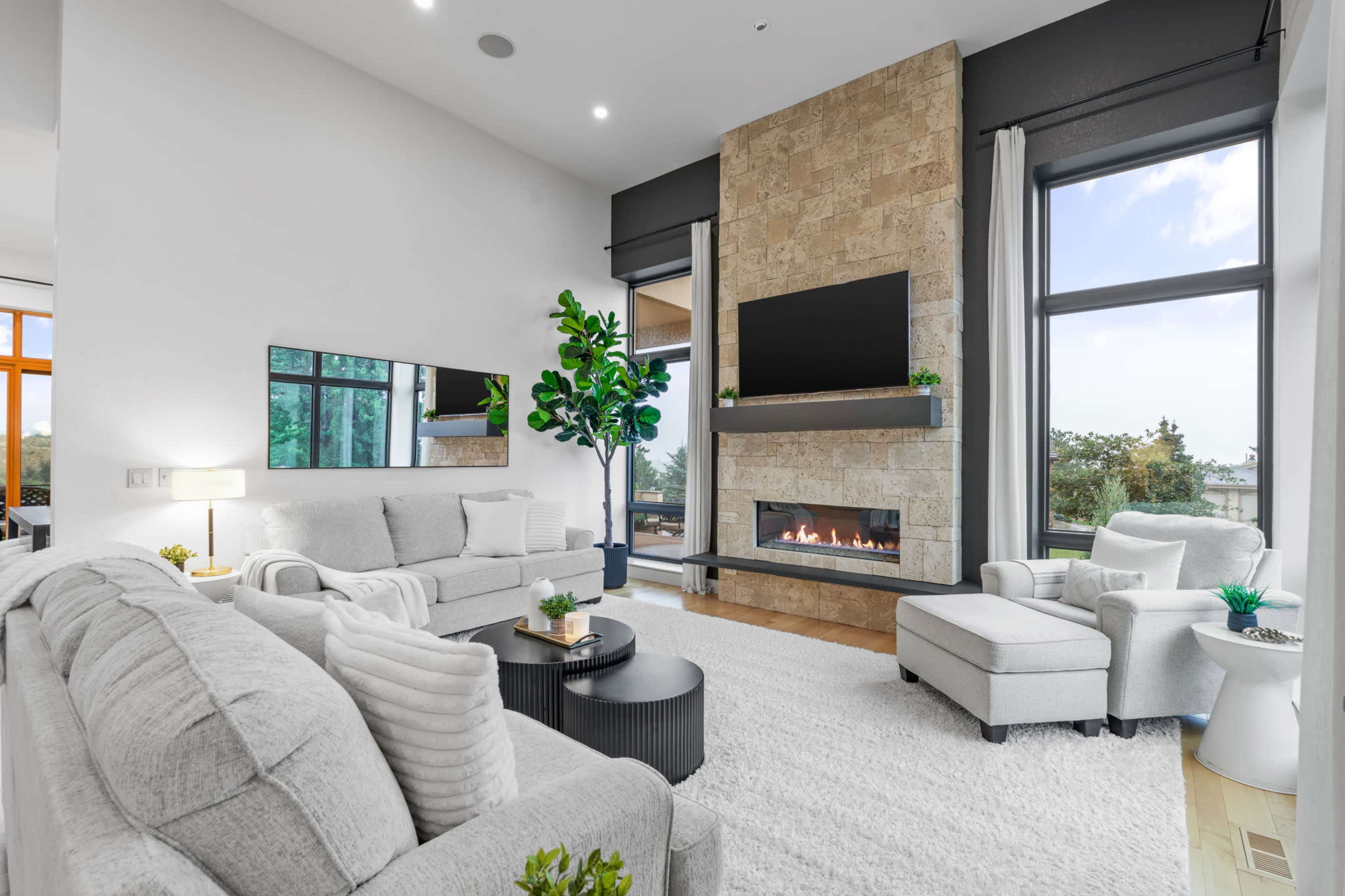 The image shows a modern living room featuring a stone fireplace, large windows, and a light-colored sofa set arranged around a coffee table.