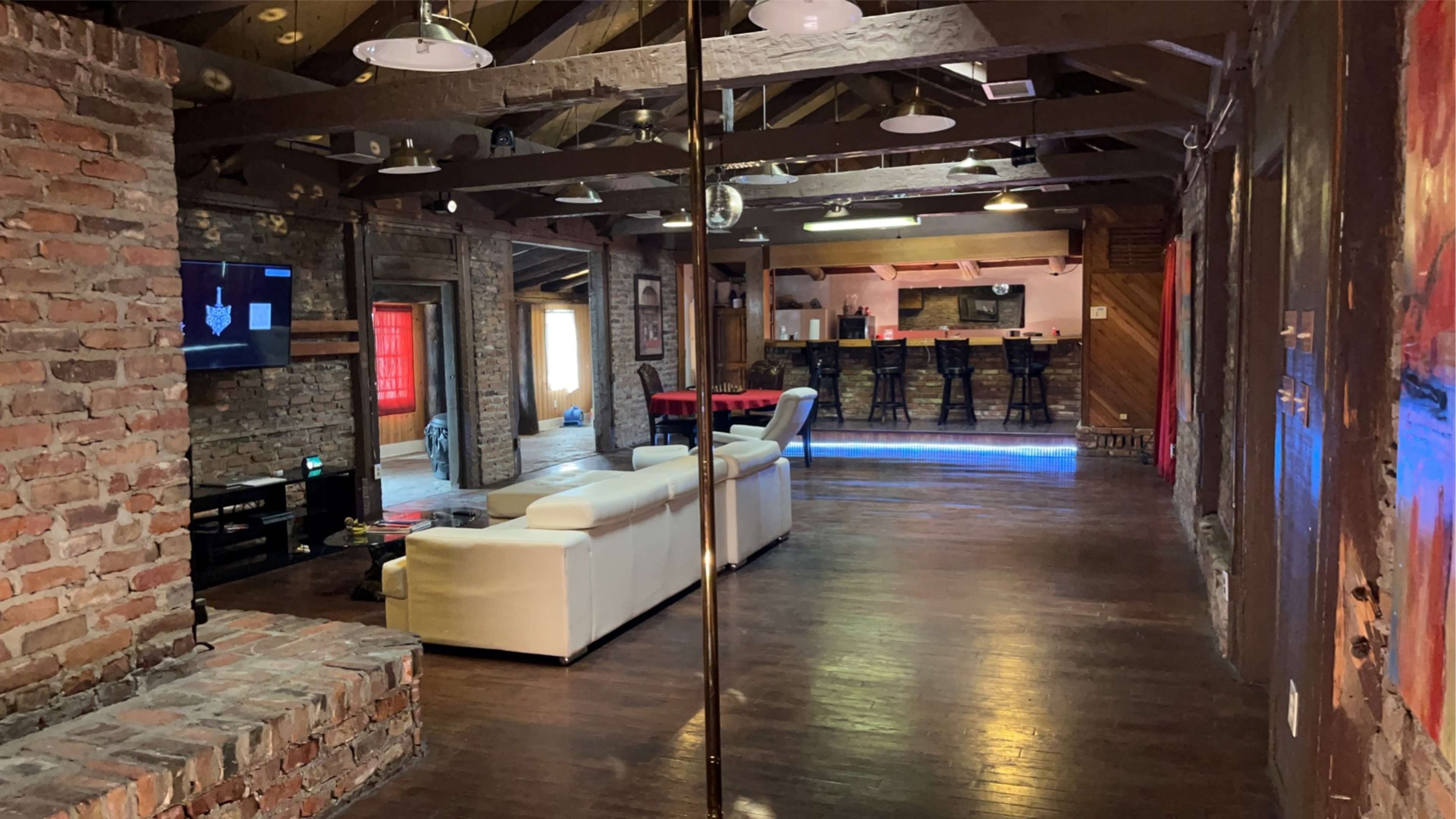 An interior space featuring a polished wooden floor, brick walls, a white sectional sofa, and a bar area with high stools in the background.