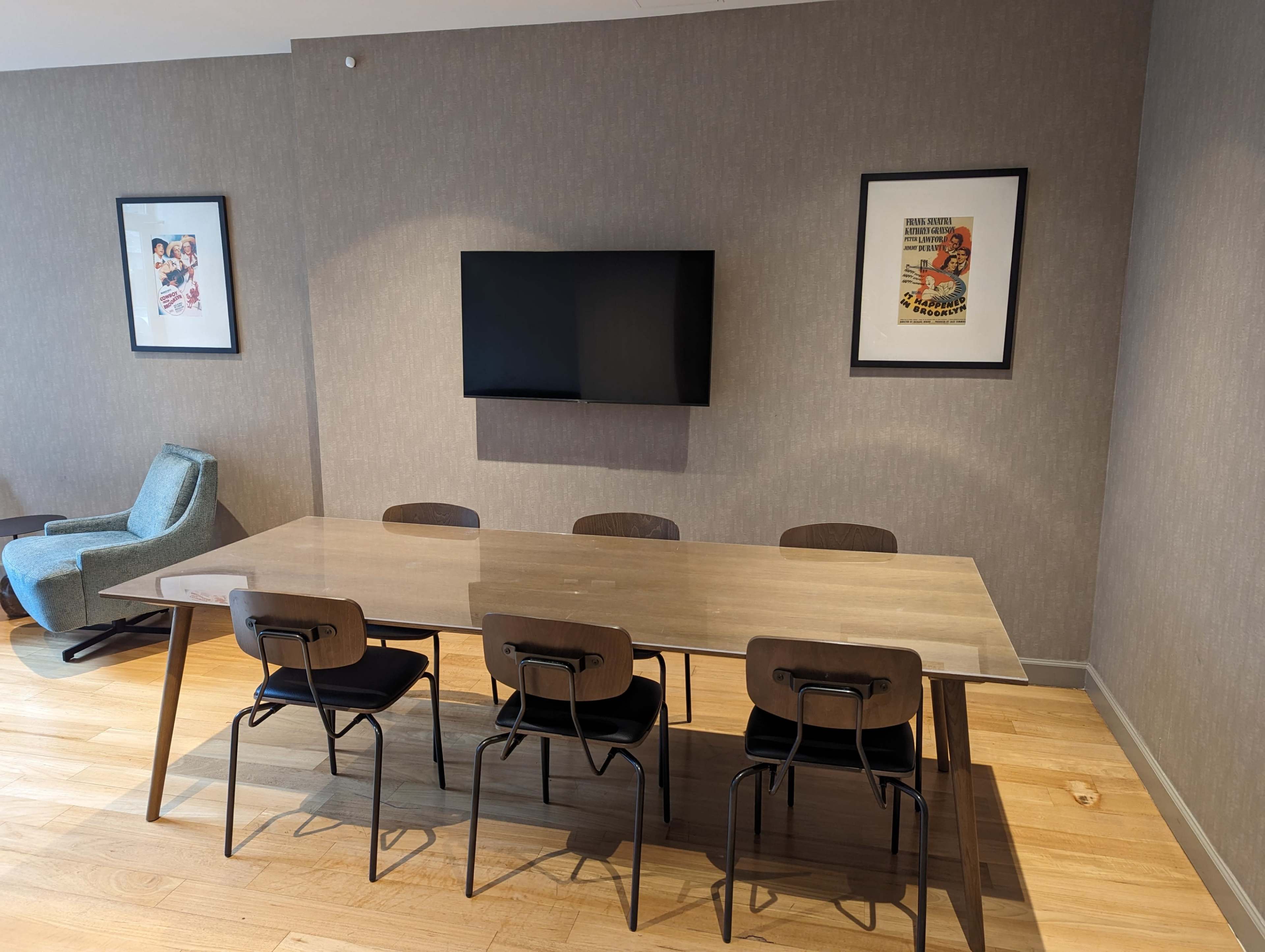 A dining area features a long table with six black chairs, a wall-mounted TV, and two framed artworks on the walls.