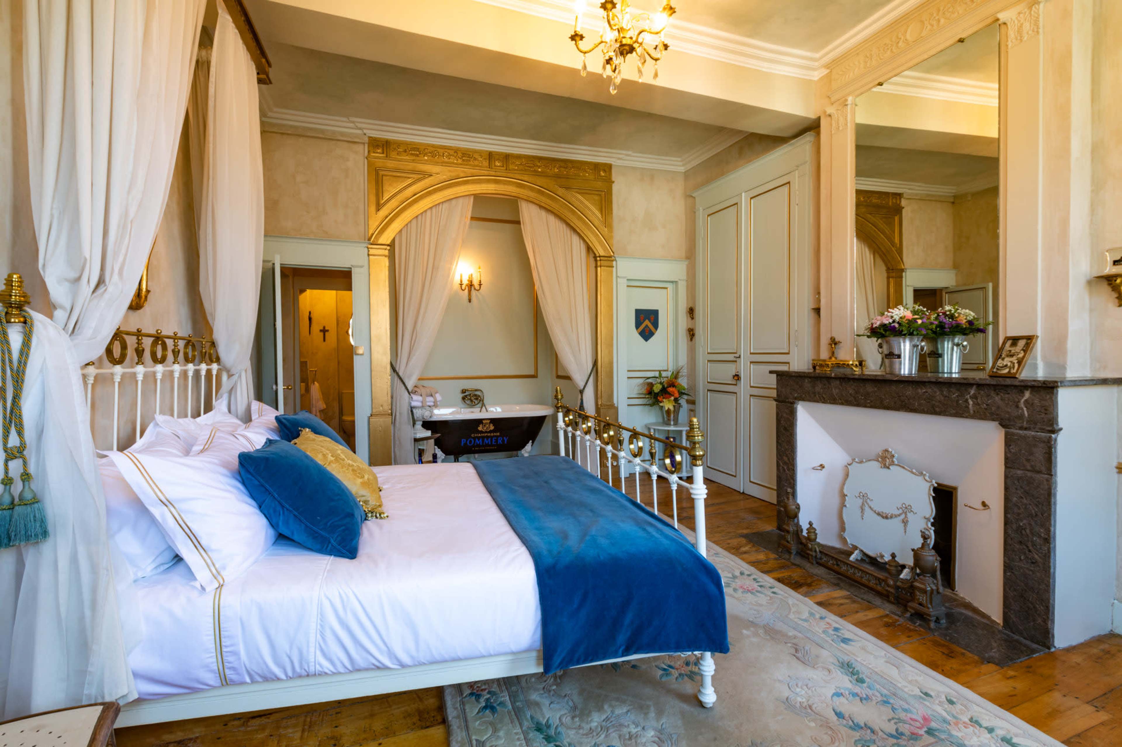 The image shows a well-appointed bedroom featuring a bed with decorative pillows, an ornate fireplace, and a large mirror reflecting the room's architecture.