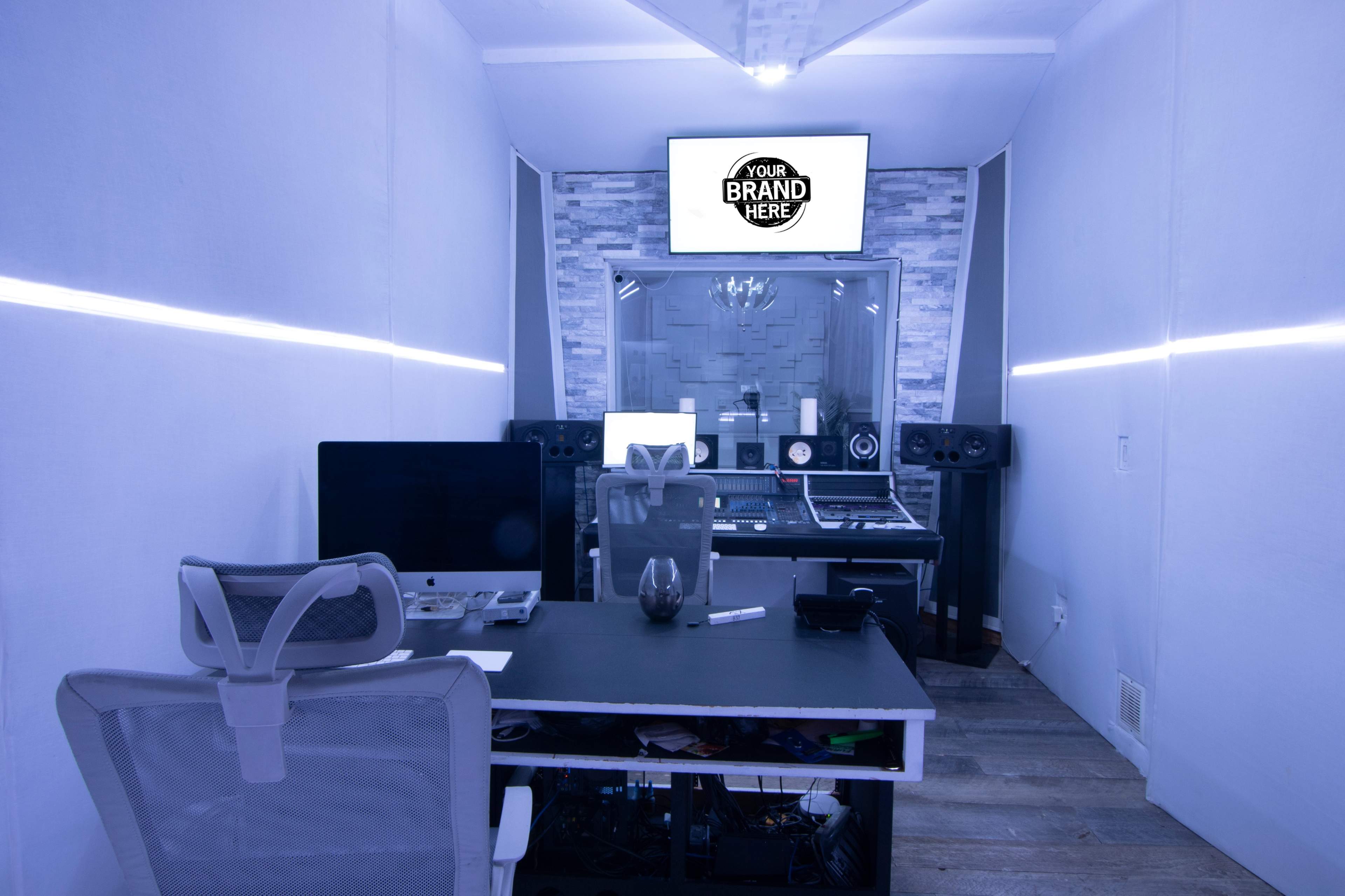 A modern recording studio features a black desk with a chair, audio equipment, and a screen displaying “YOUR BRAND HERE” on the wall.