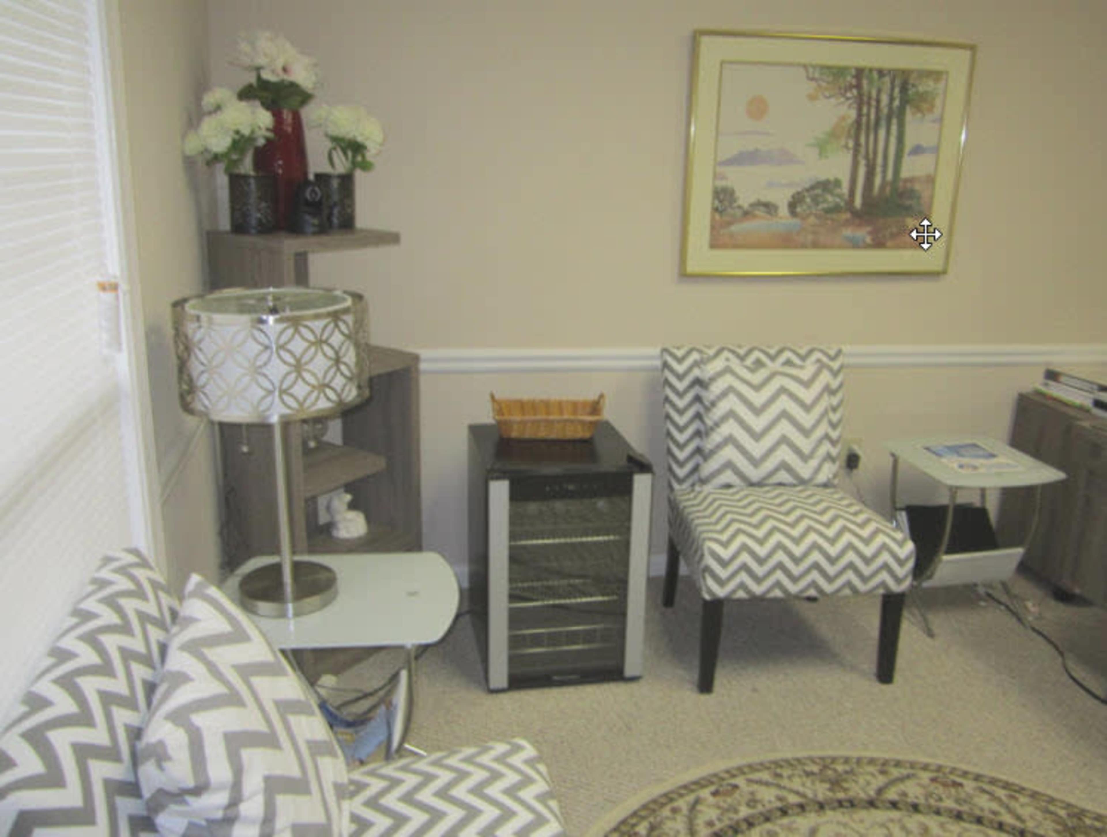 The image shows a small living area featuring two patterned chairs, a side table with a lamp, a wine cooler, and a decorative wall art piece.
