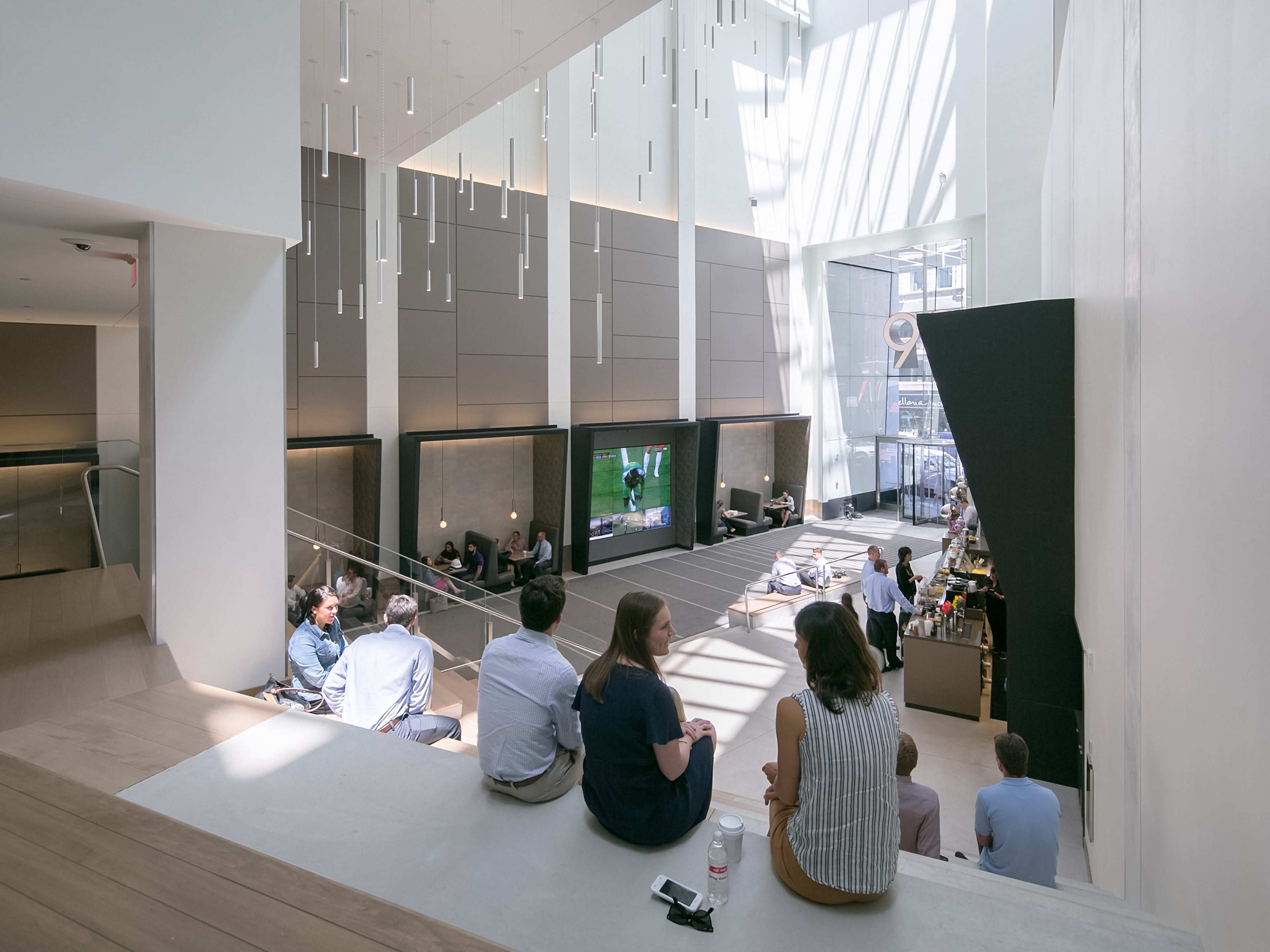 A modern interior space with a large open area, featuring a group of people seated on a staircase and a screen displaying a sports event.