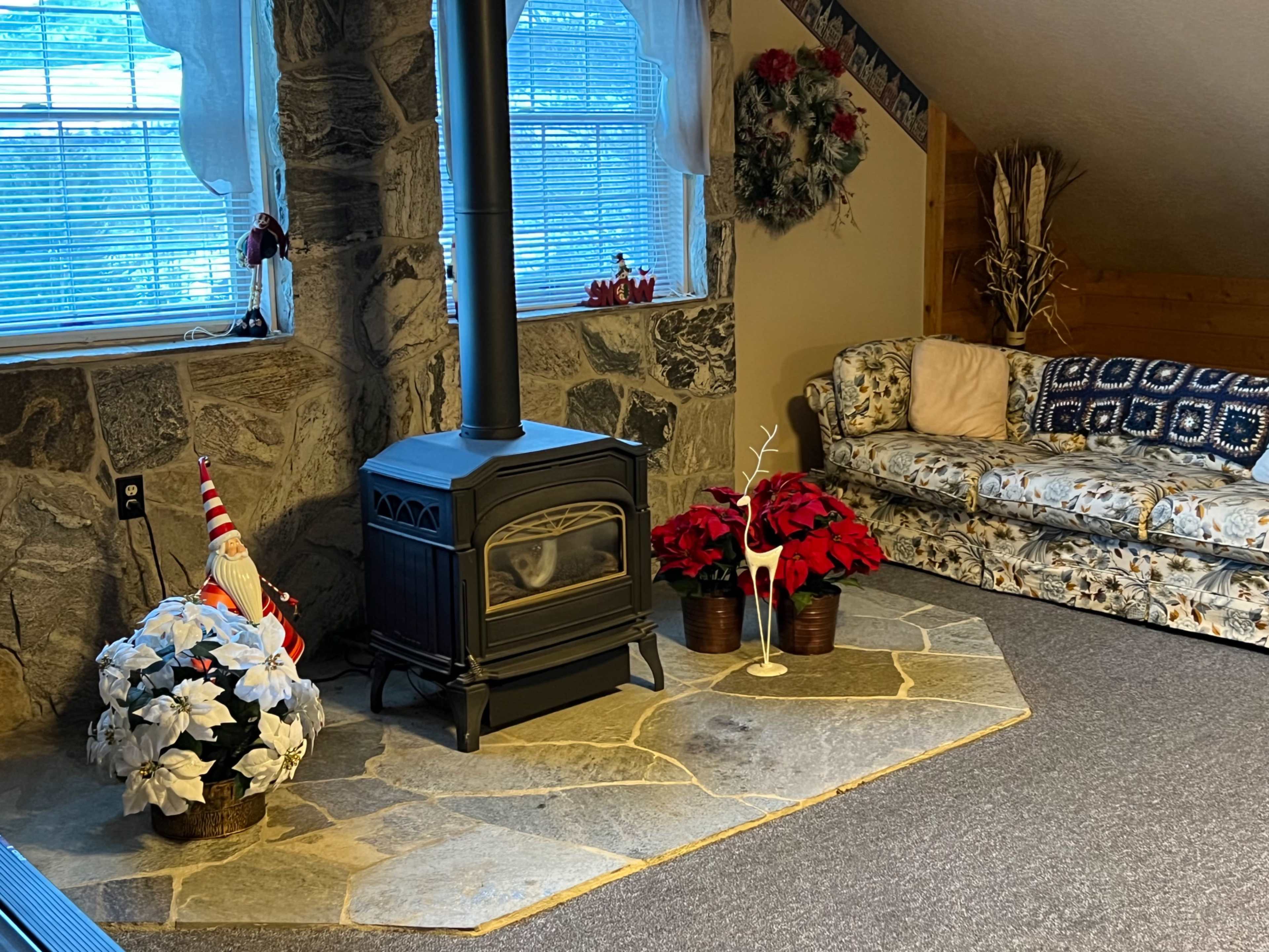 The image shows a living room with a stone fireplace, a wood stove, holiday decorations, and a floral-patterned couch.