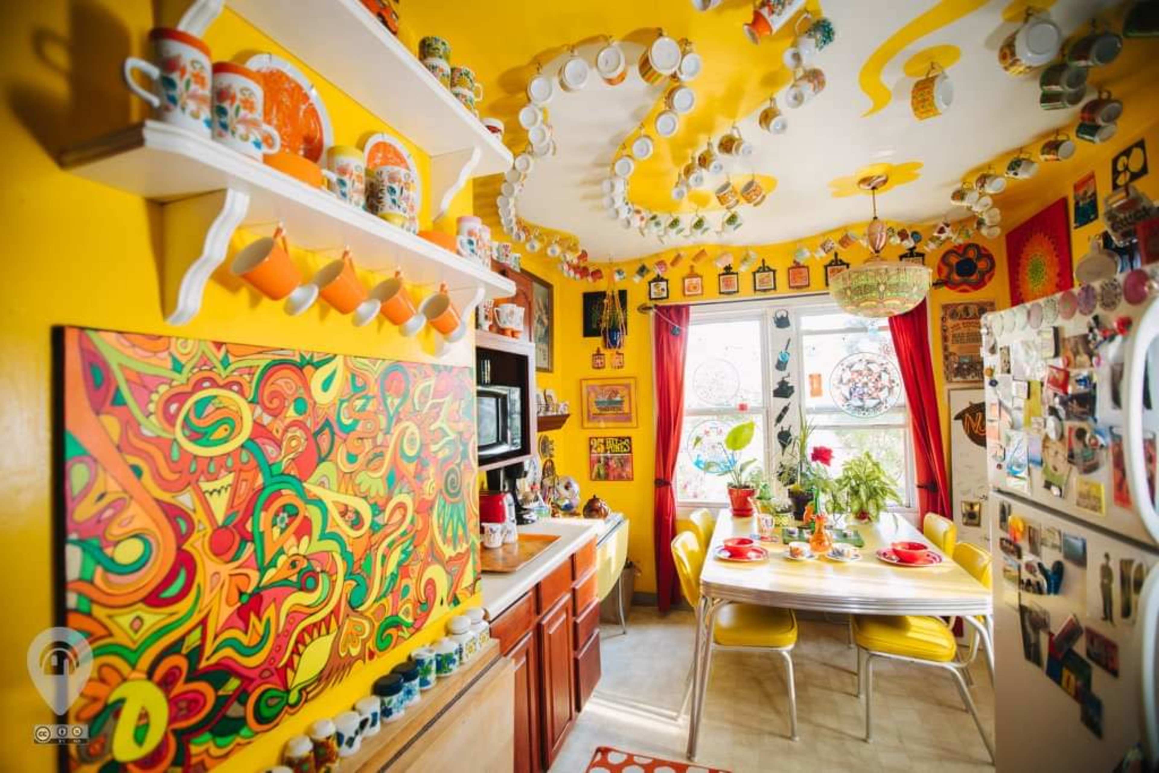 The kitchen features bright yellow walls adorned with colorful artwork and decorative cups, a table set for a meal, and a variety of kitchenware displayed on shelves.
