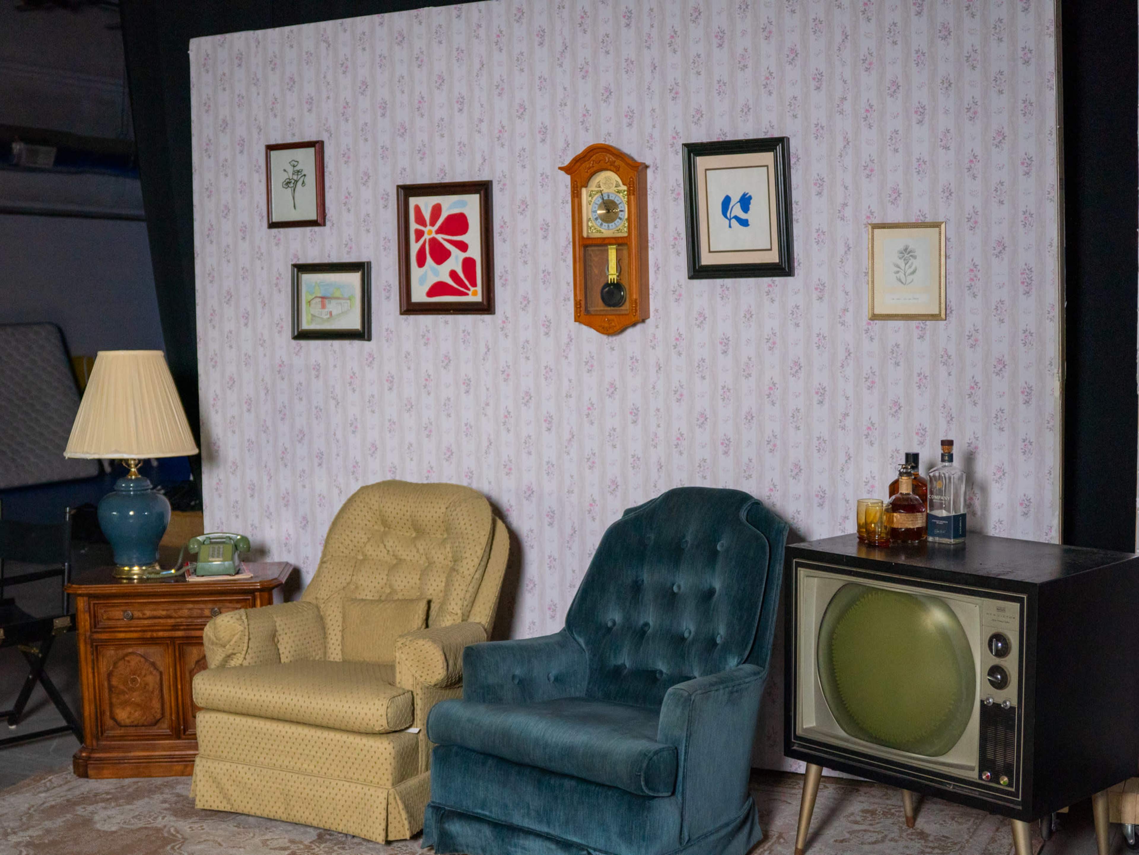 The image features a vintage living room setup with two armchairs, a side table, a wall clock, framed artworks, and an old-fashioned television, all against a patterned wallpaper backdrop.