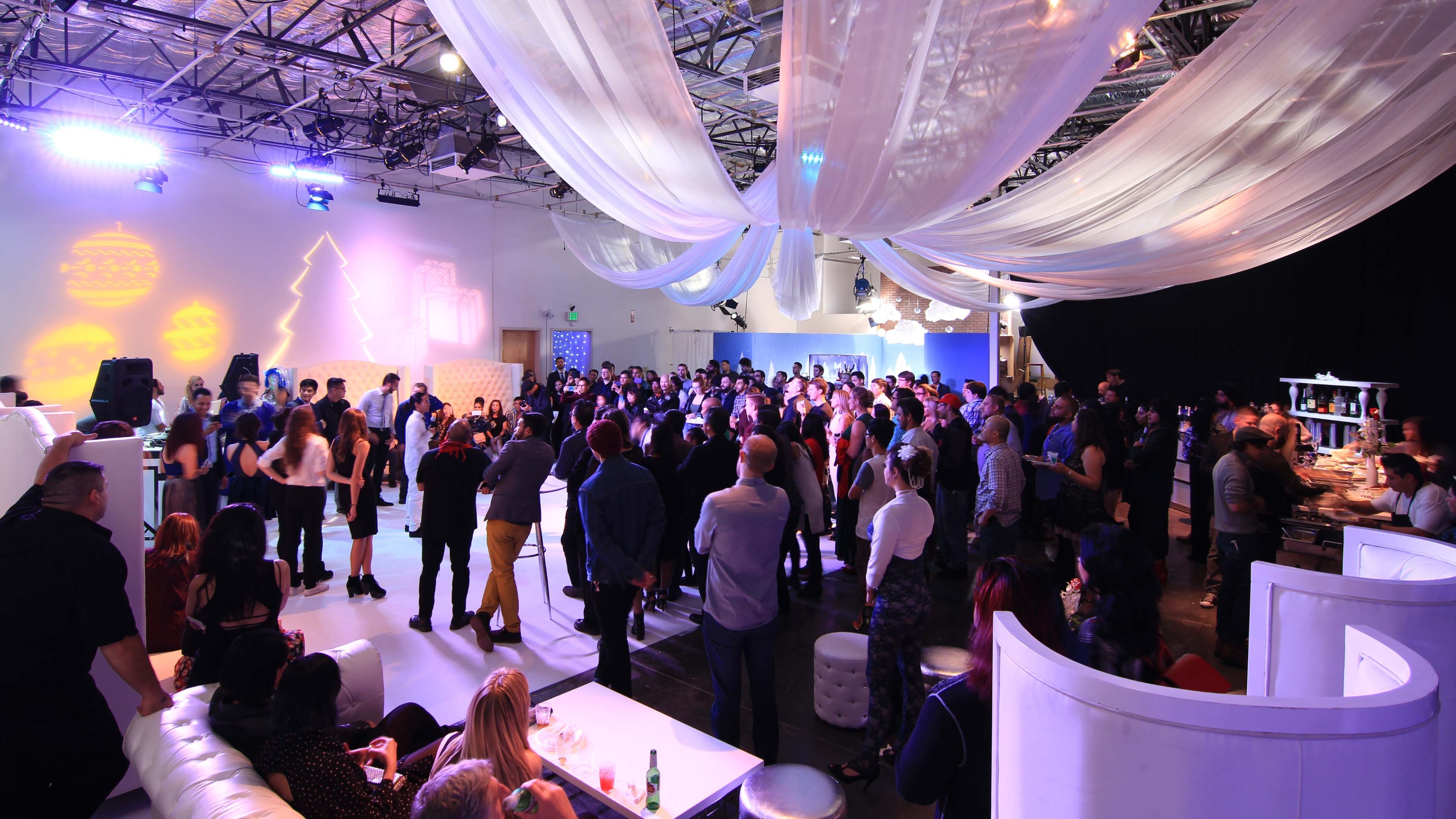 A large group of people gathers in a spacious venue decorated with draped fabric and festive lighting.