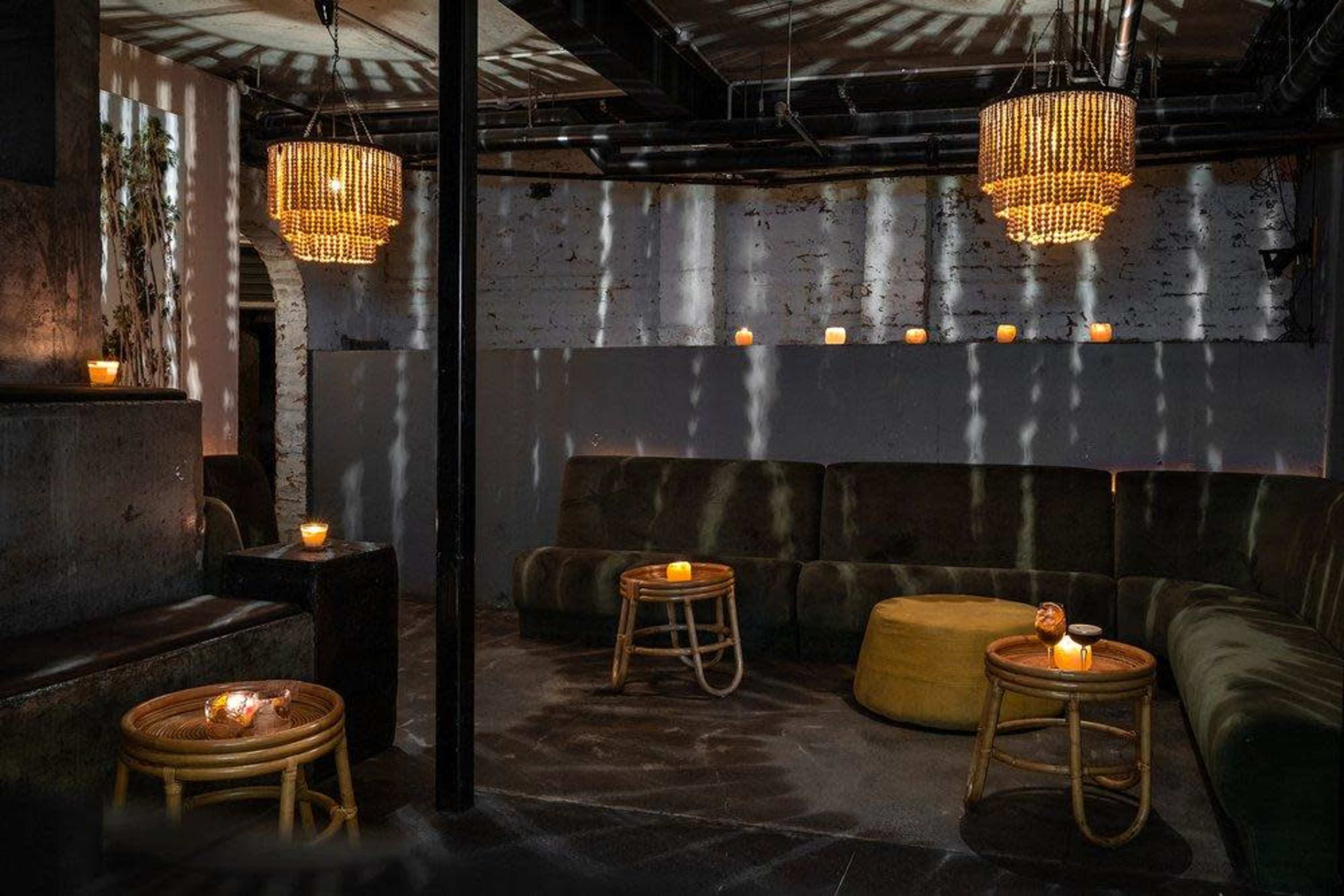 A dimly lit lounge area featuring plush seating, small wooden tables, and candlelight casting shadows on the walls.
