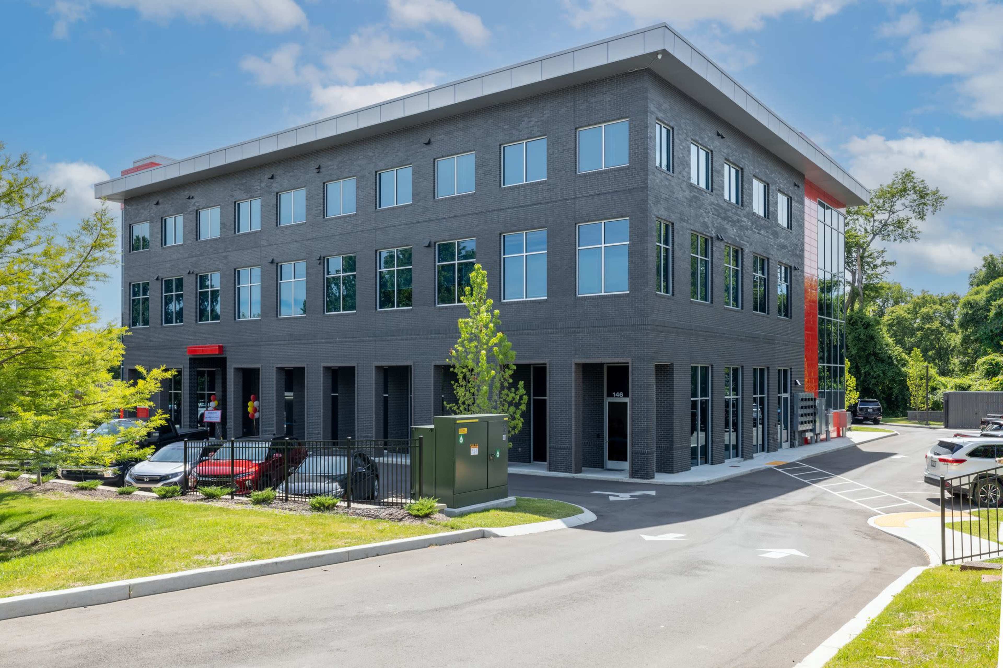 A modern, three-story office building featuring a mix of gray brick and large windows, surrounded by landscaped grounds and a parking area.