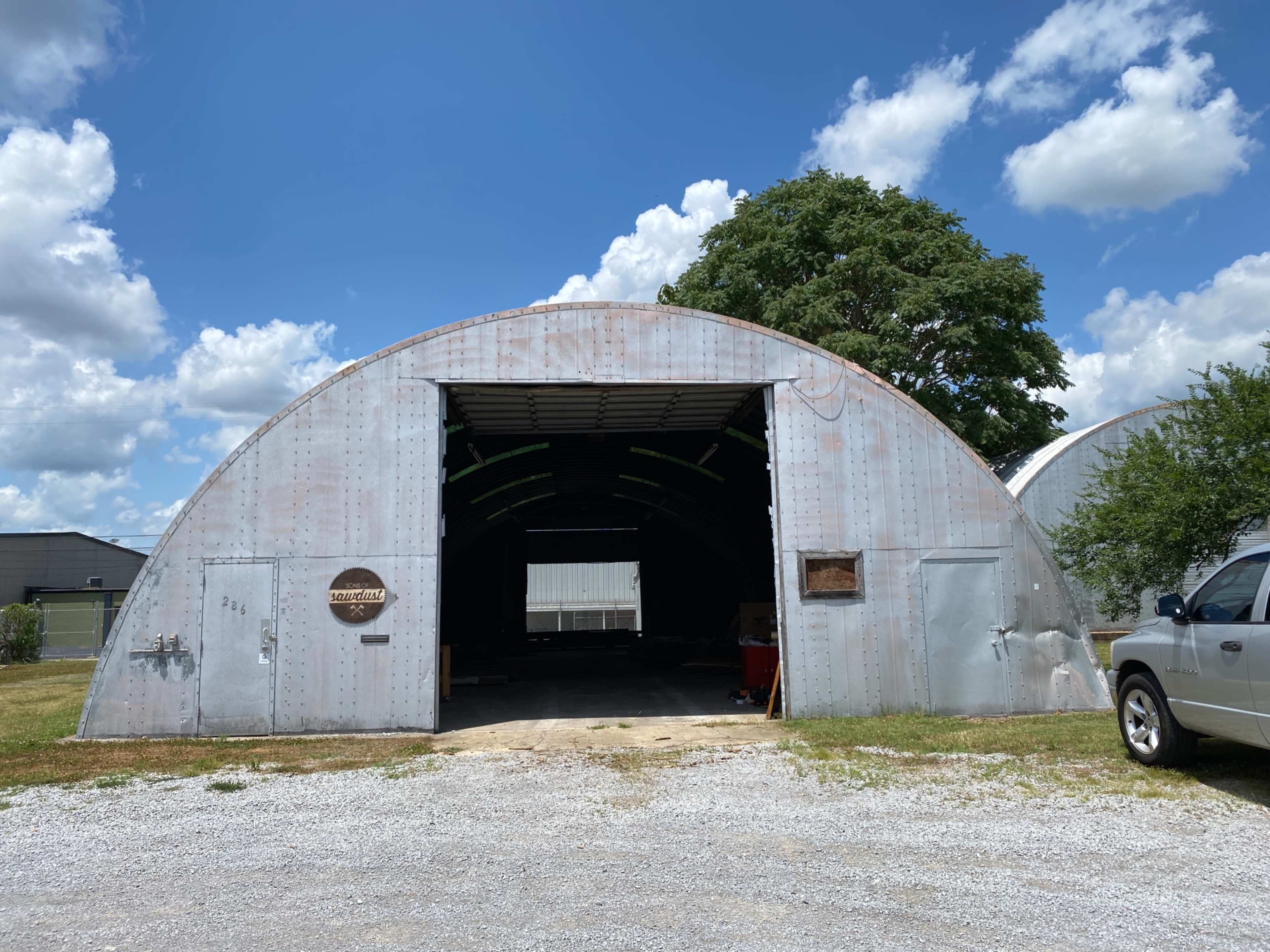 A large, corrugated metal structure with an arched roof and an open entrance, surrounded by gravel and green vegetation under a cloudy blue sky.