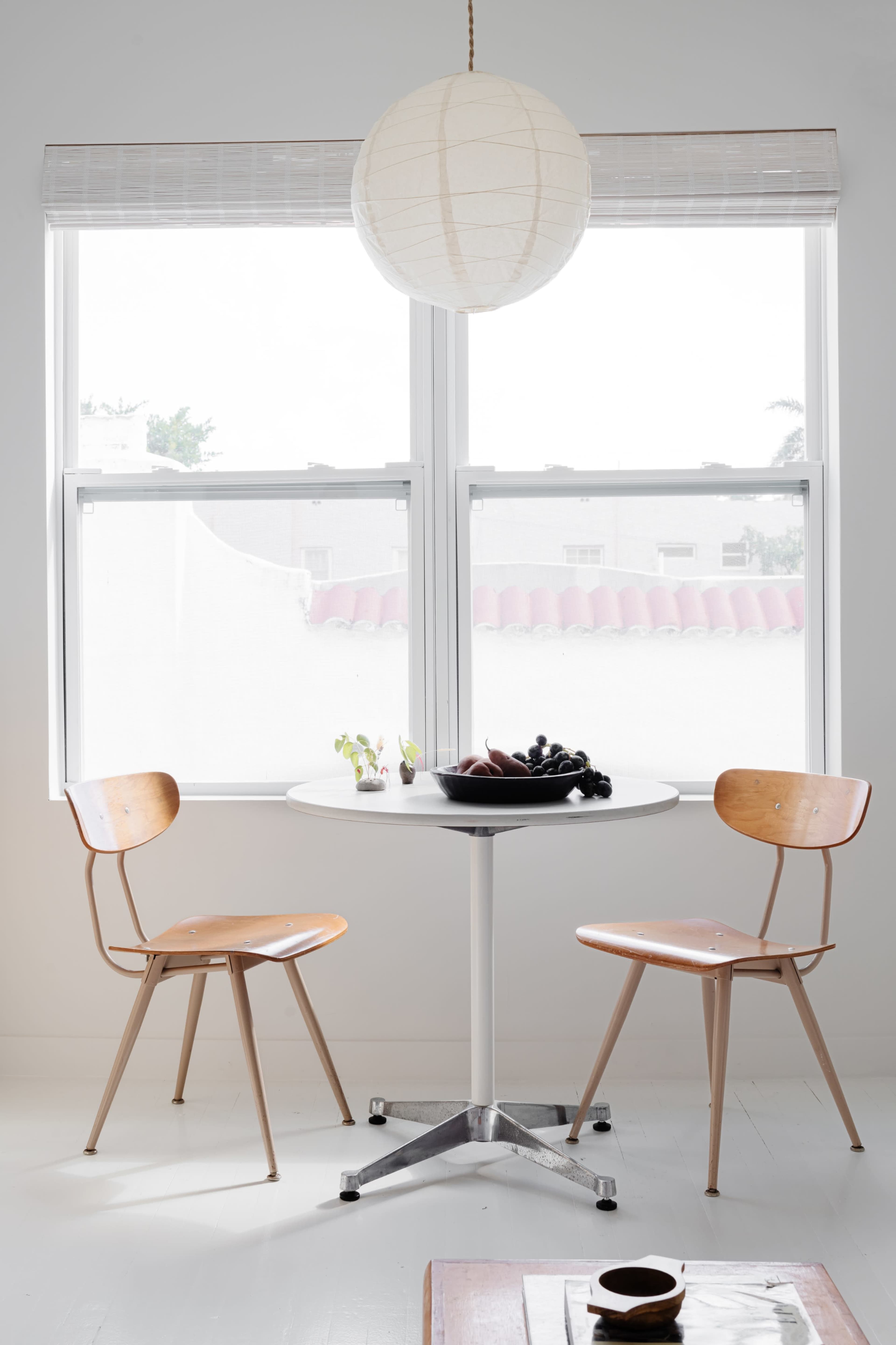 A small round table with a fruit arrangement is positioned between two wooden chairs in a bright room with large windows.