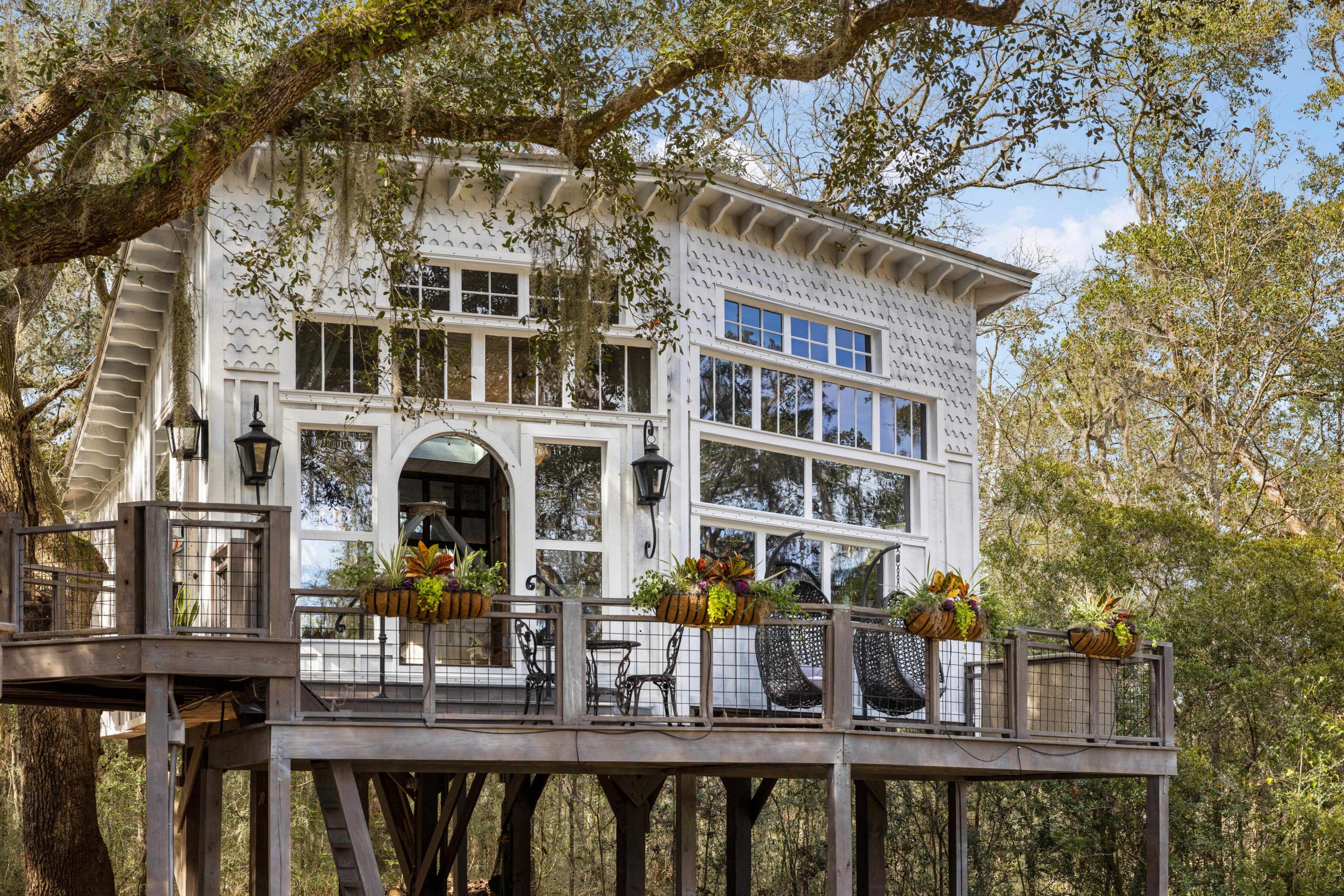 A elevated, wooden house features large windows, a spacious deck with seating, and is surrounded by trees.
