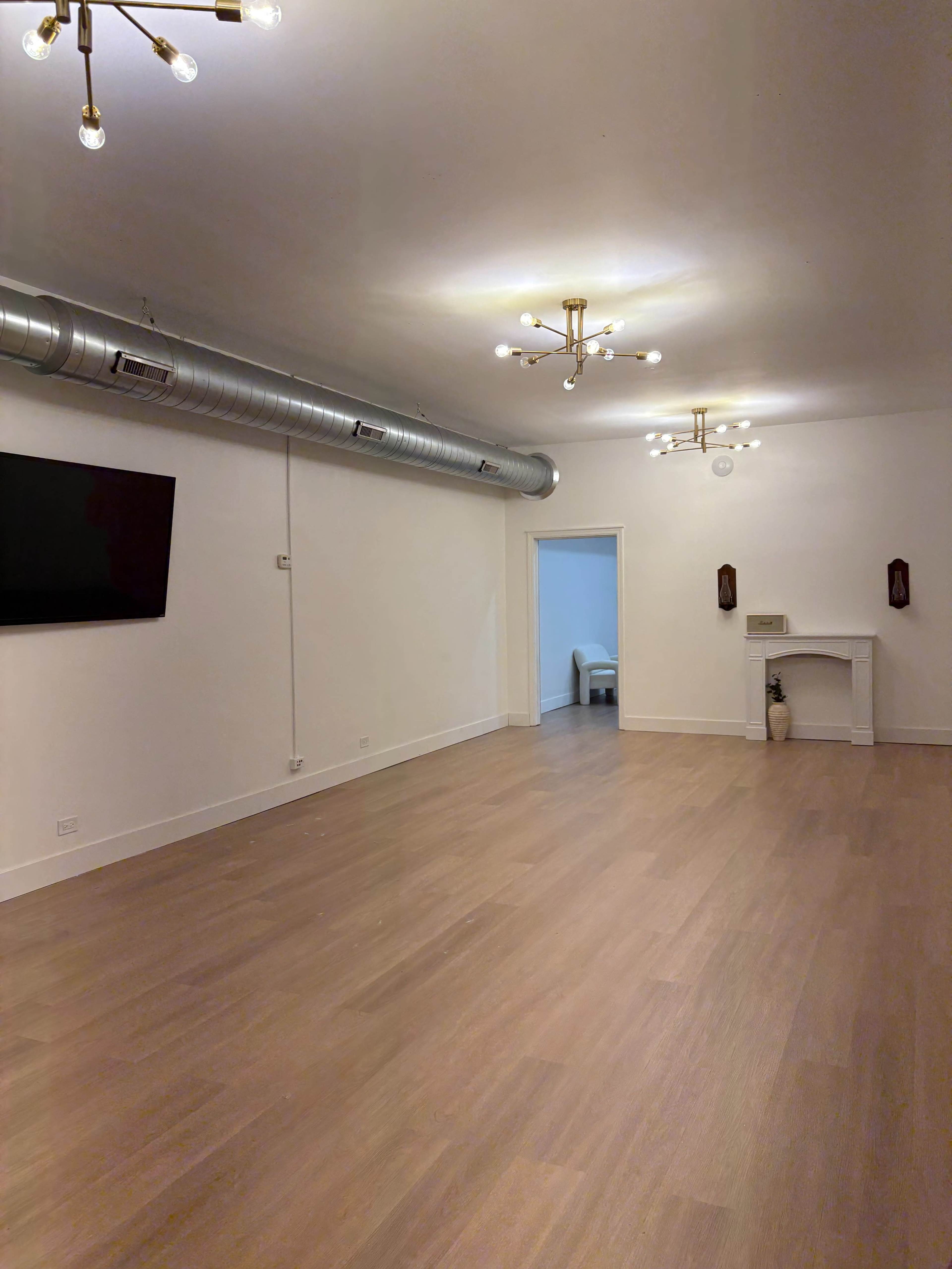 The image shows a spacious, empty room with wooden flooring, overhead lighting fixtures, and a small fireplace against one wall.