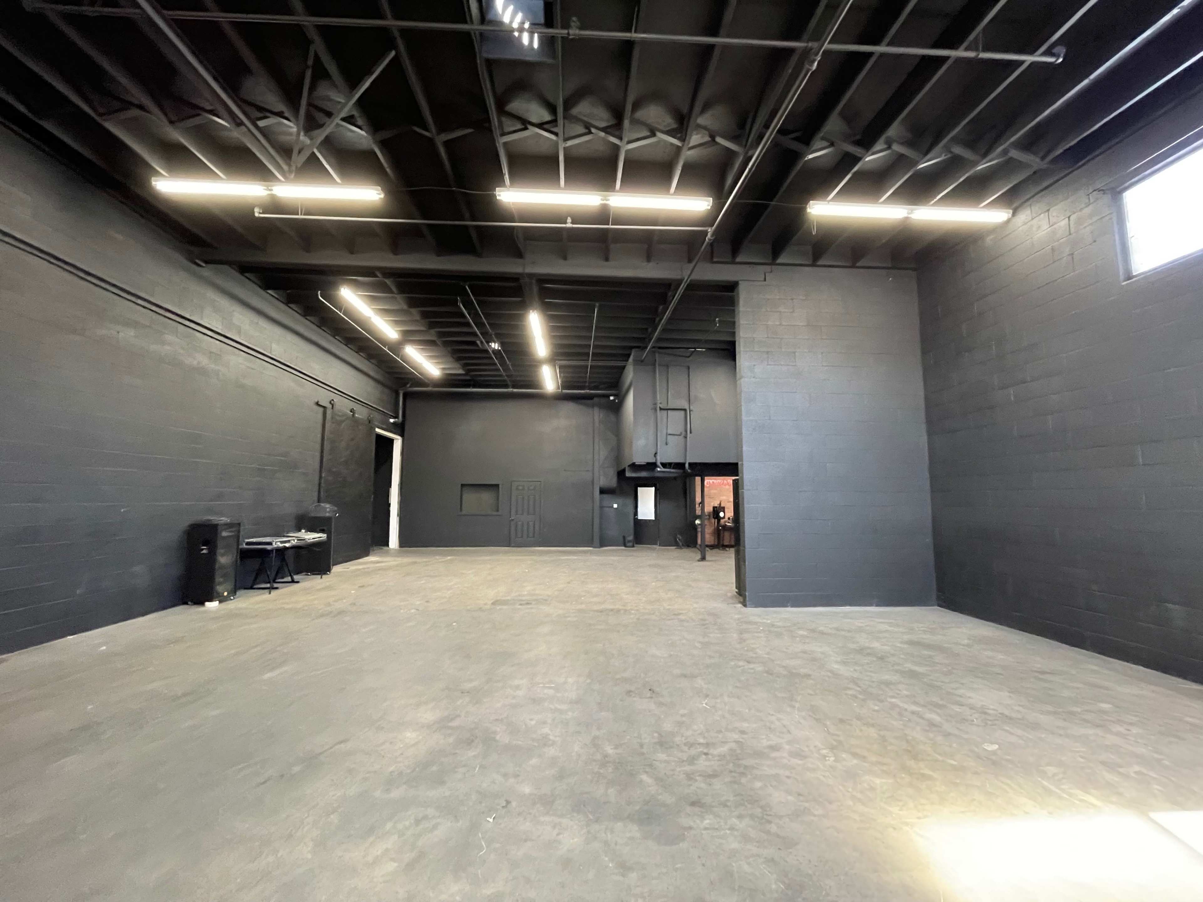 The image shows a spacious, industrial-style room with black walls and a concrete floor, featuring overhead lighting and a large open area.