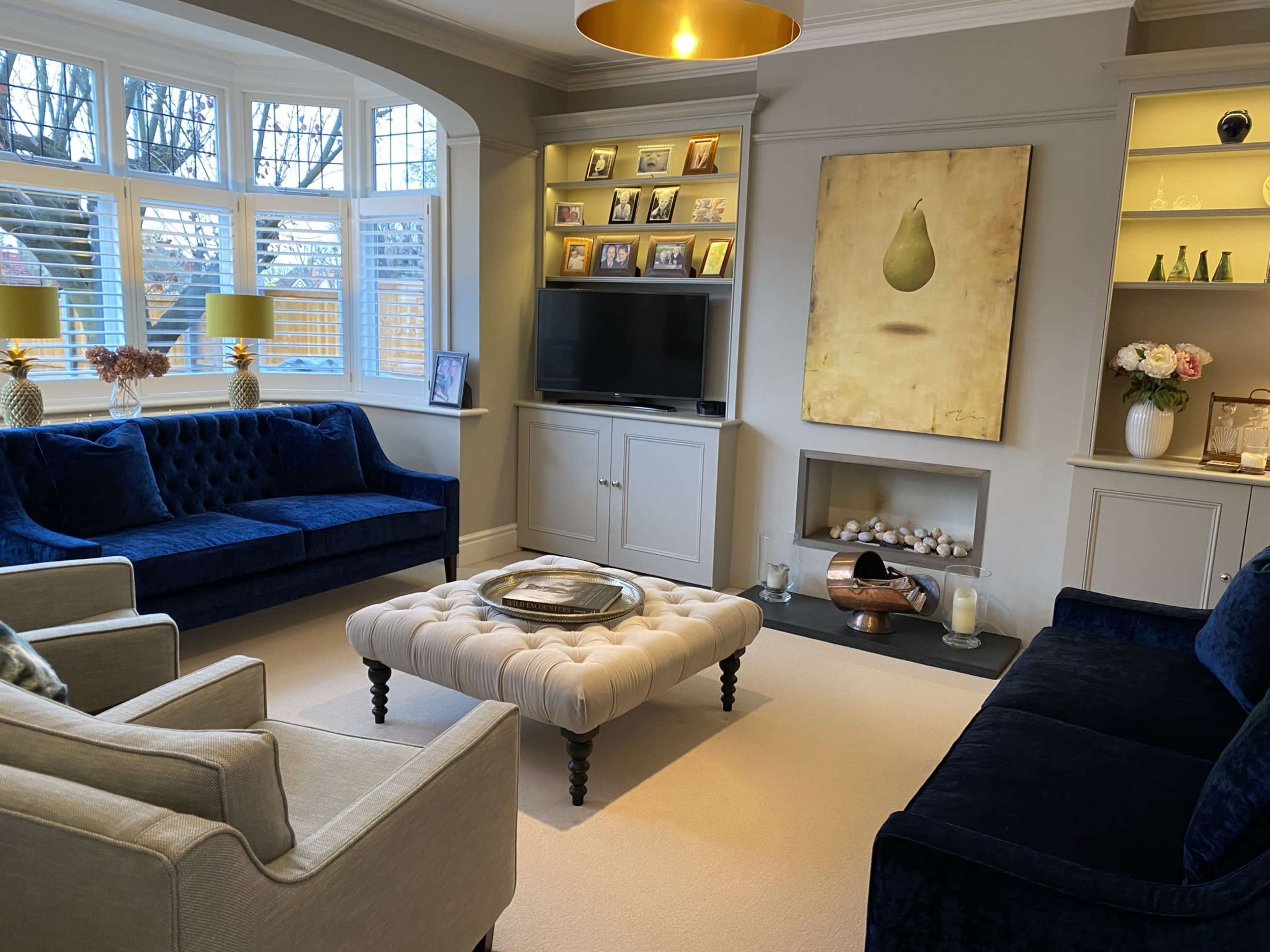 A modern living room features two blue velvet sofas, a tufted ottoman, a television, and a cream-colored area rug, with a wall art piece and shelves displaying photos and decorative items.