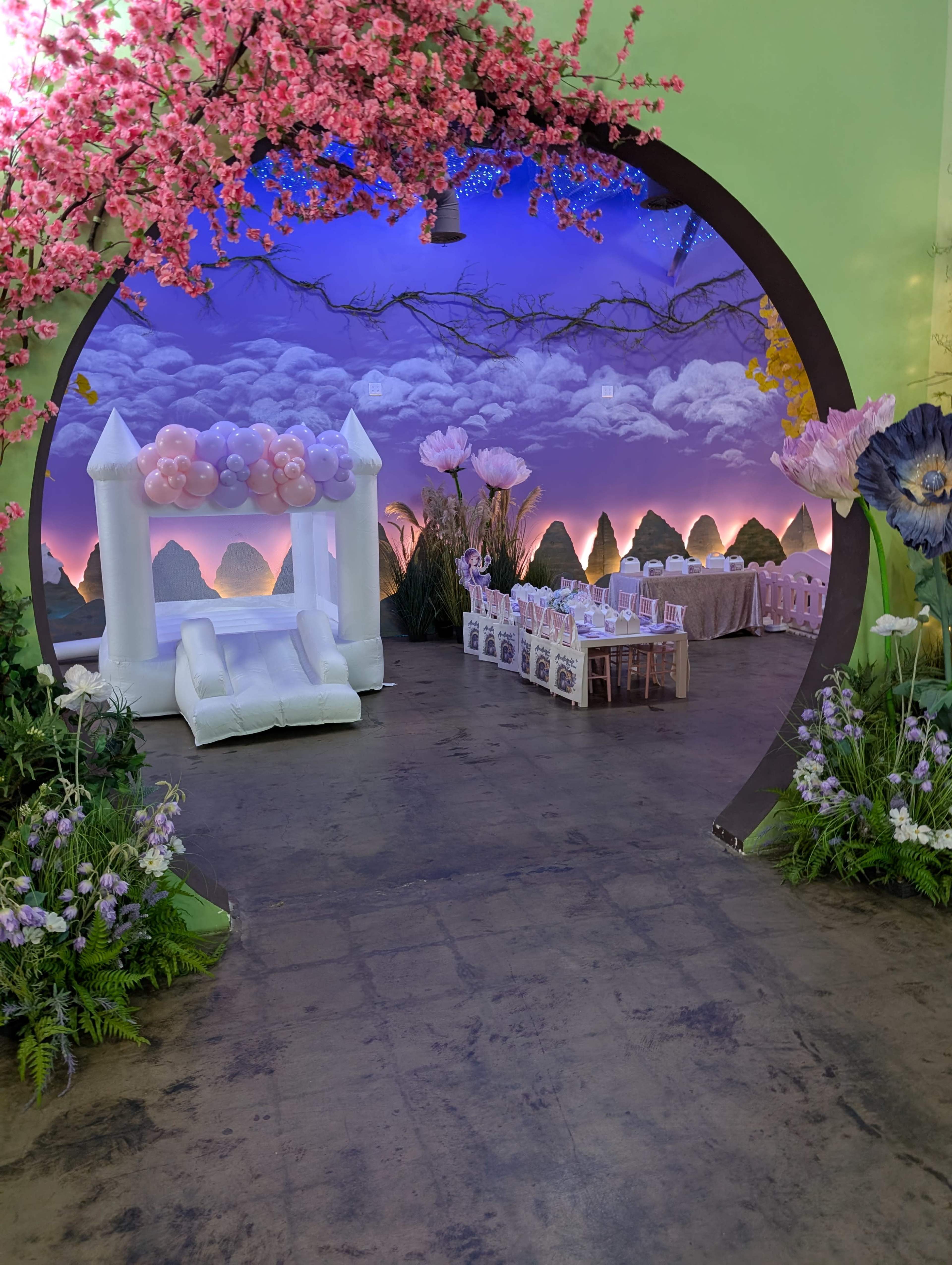 The image shows a party space adorned with pink blossoms, featuring a bounce house and a table set for a celebration.