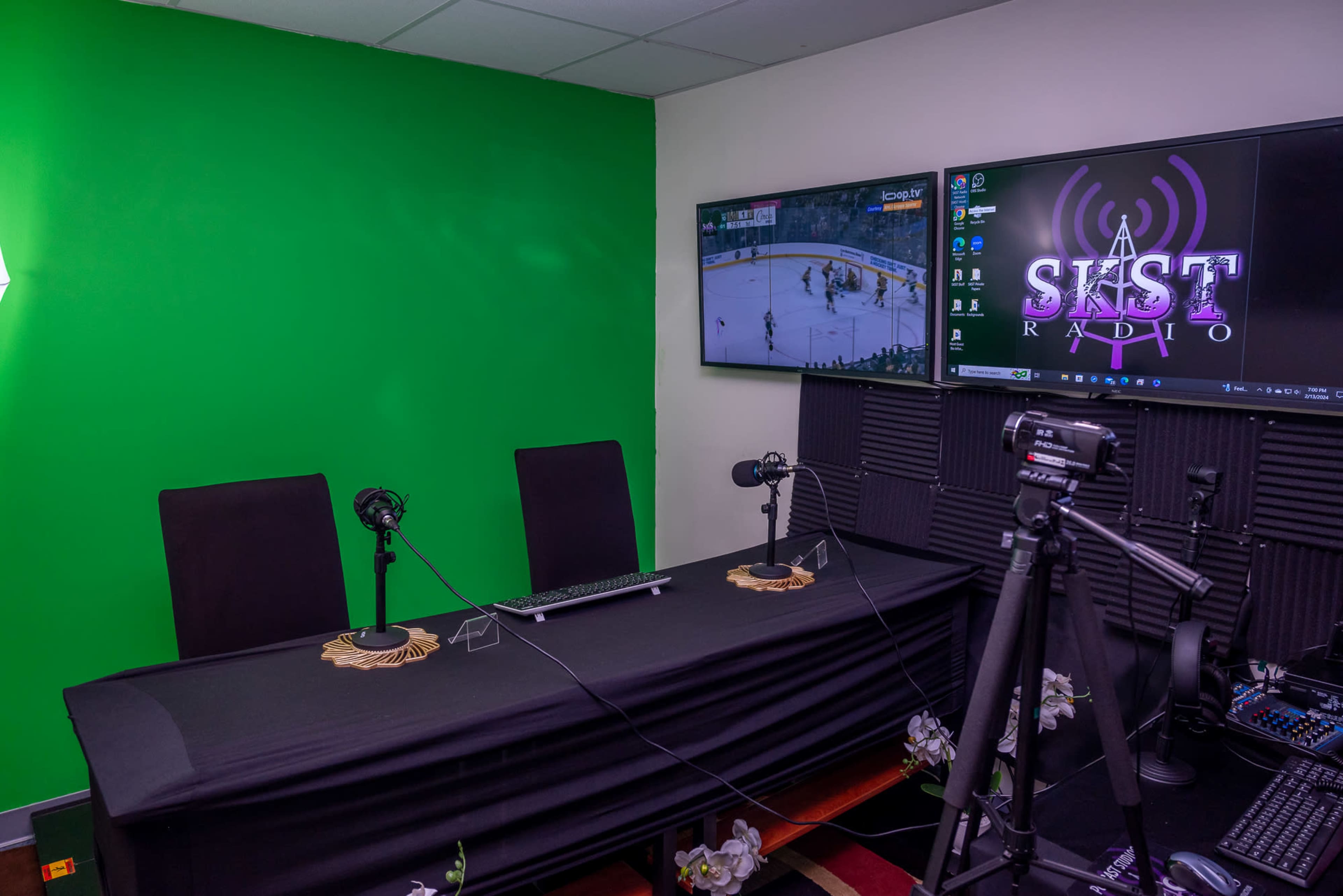 A recording studio with a green screen, two microphones, a desktop computer, and televisions displaying a sports game and a logo for a radio station.