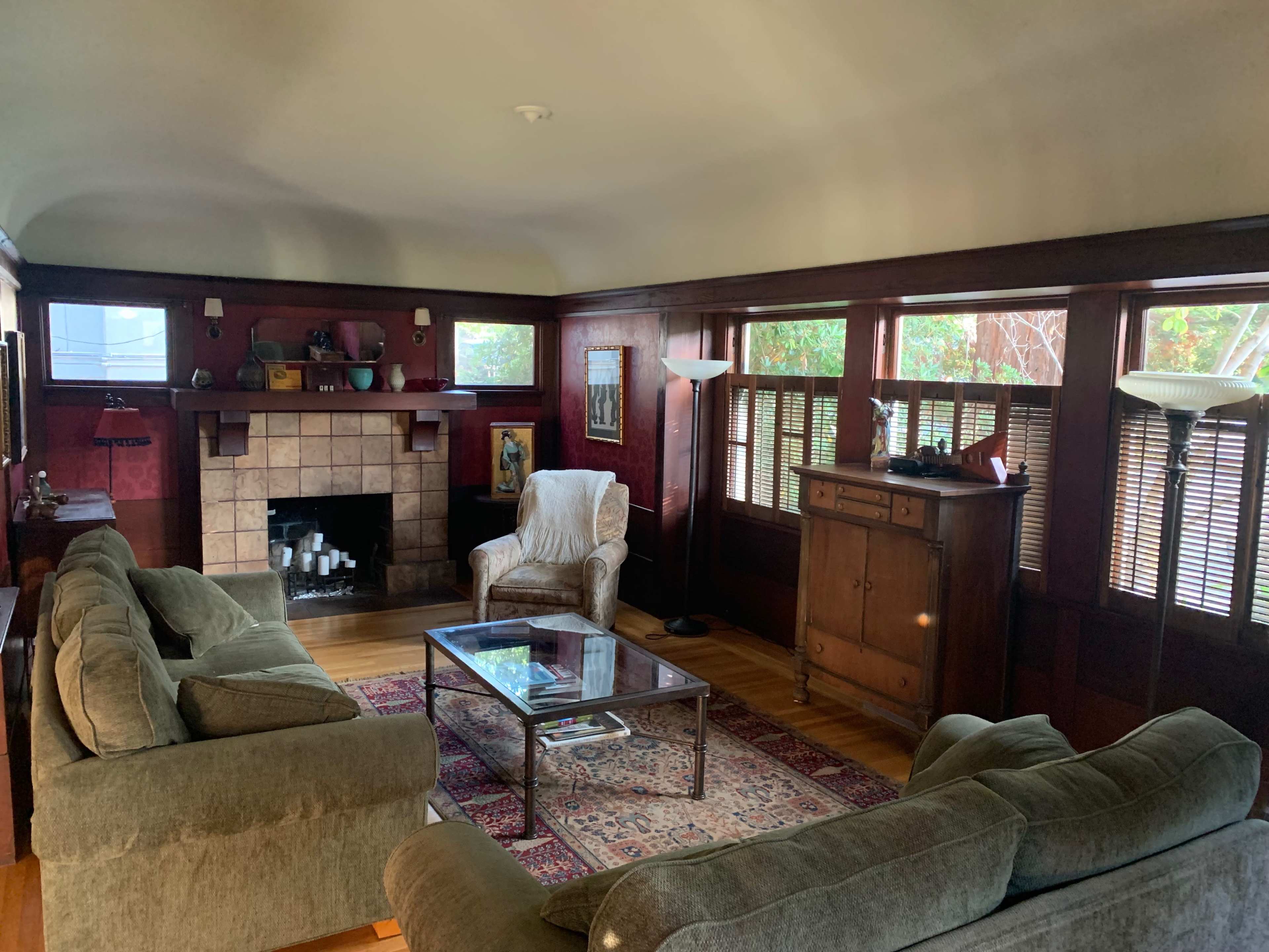 A living room features a stone fireplace, two green sofas, a coffee table, and large windows with wooden blinds.
