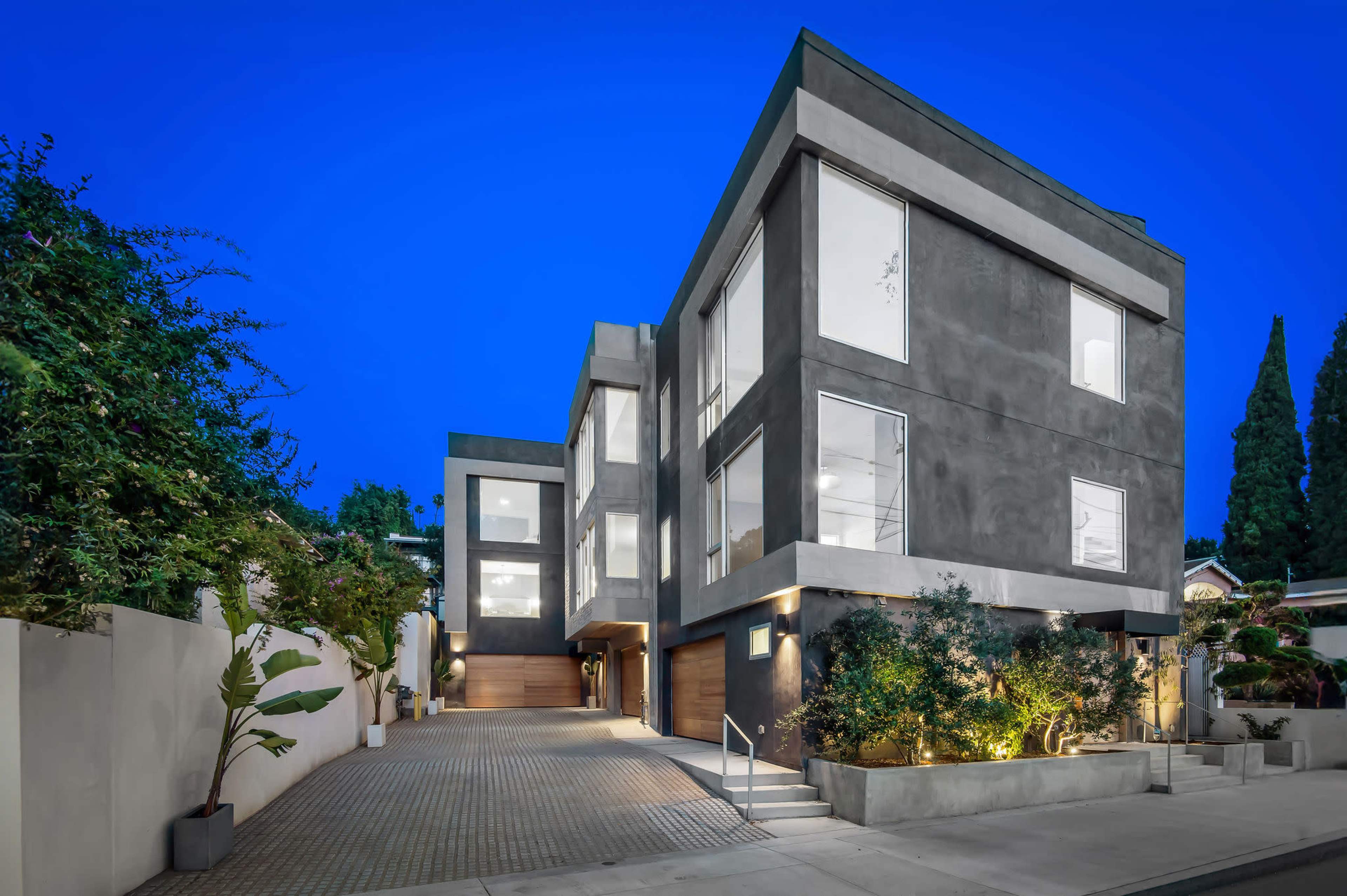 The image shows a modern, multi-story house with large windows, situated on a tree-lined street at dusk.