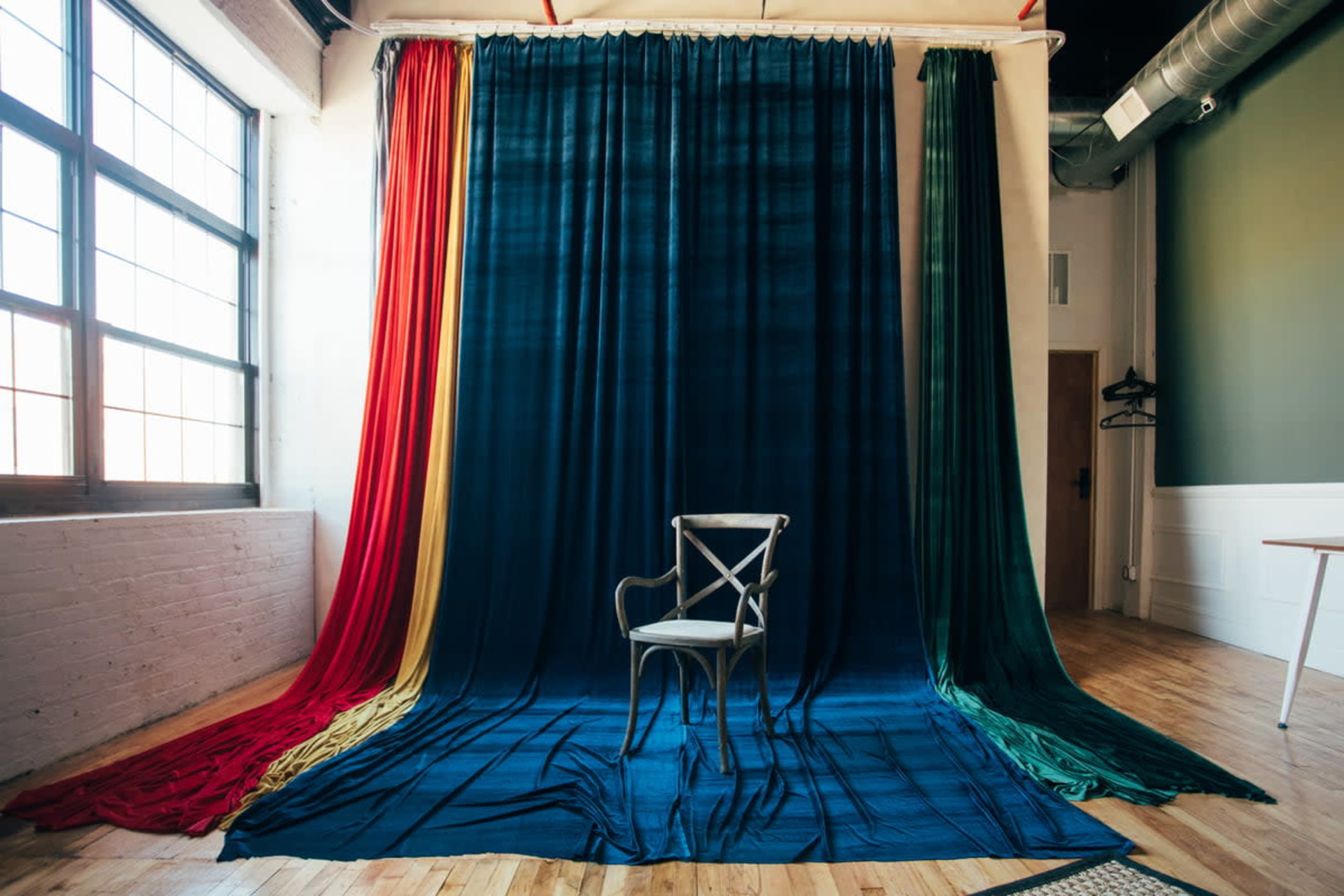 The image shows a simple wooden chair placed in front of a backdrop with draped fabric in various colors, including red, green, and dark blue, inside a well-lit studio with large windows.