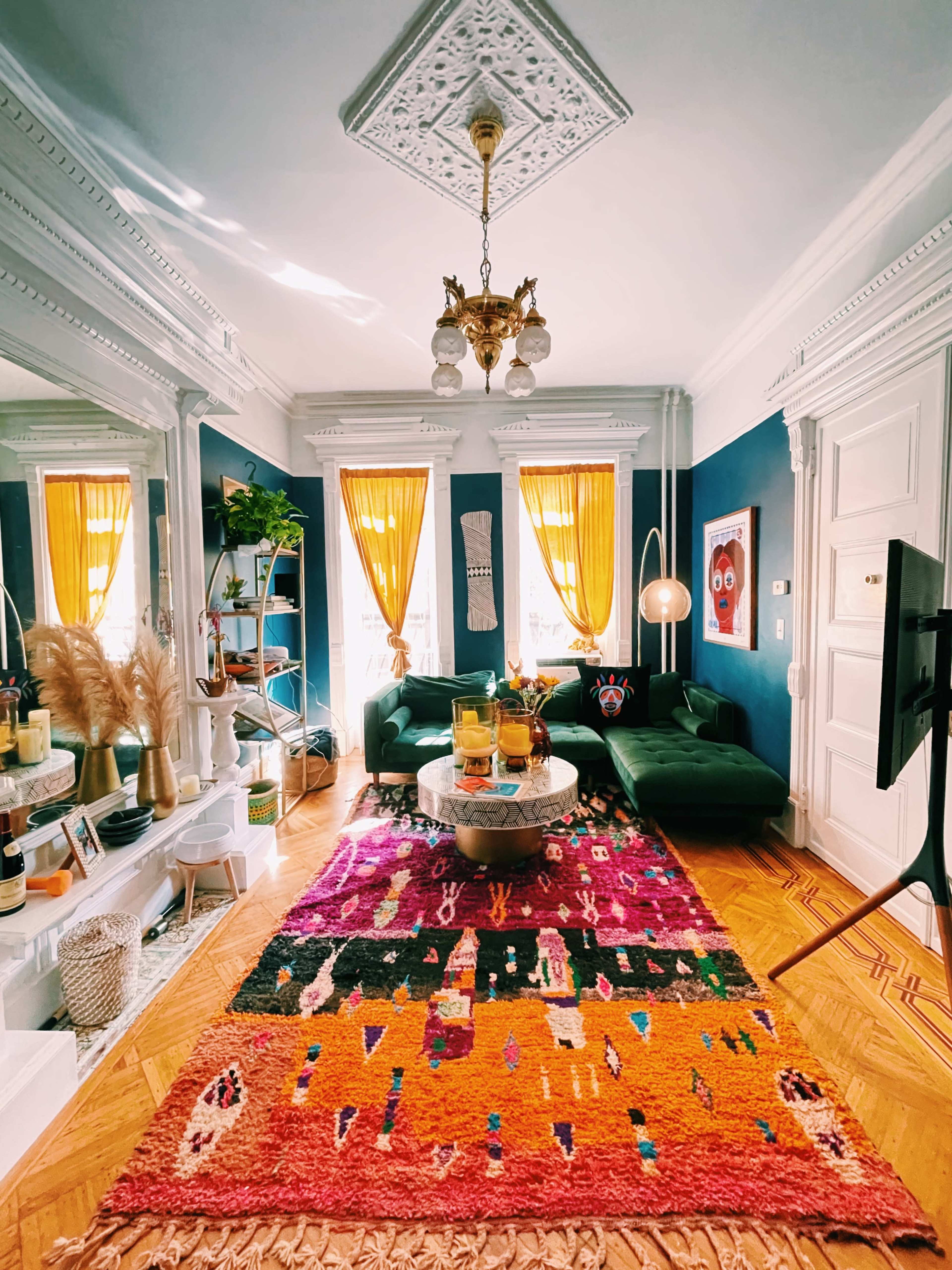 A brightly lit living room features a patterned rug, a green sofa, and decorated walls with mirrors and yellow curtains.