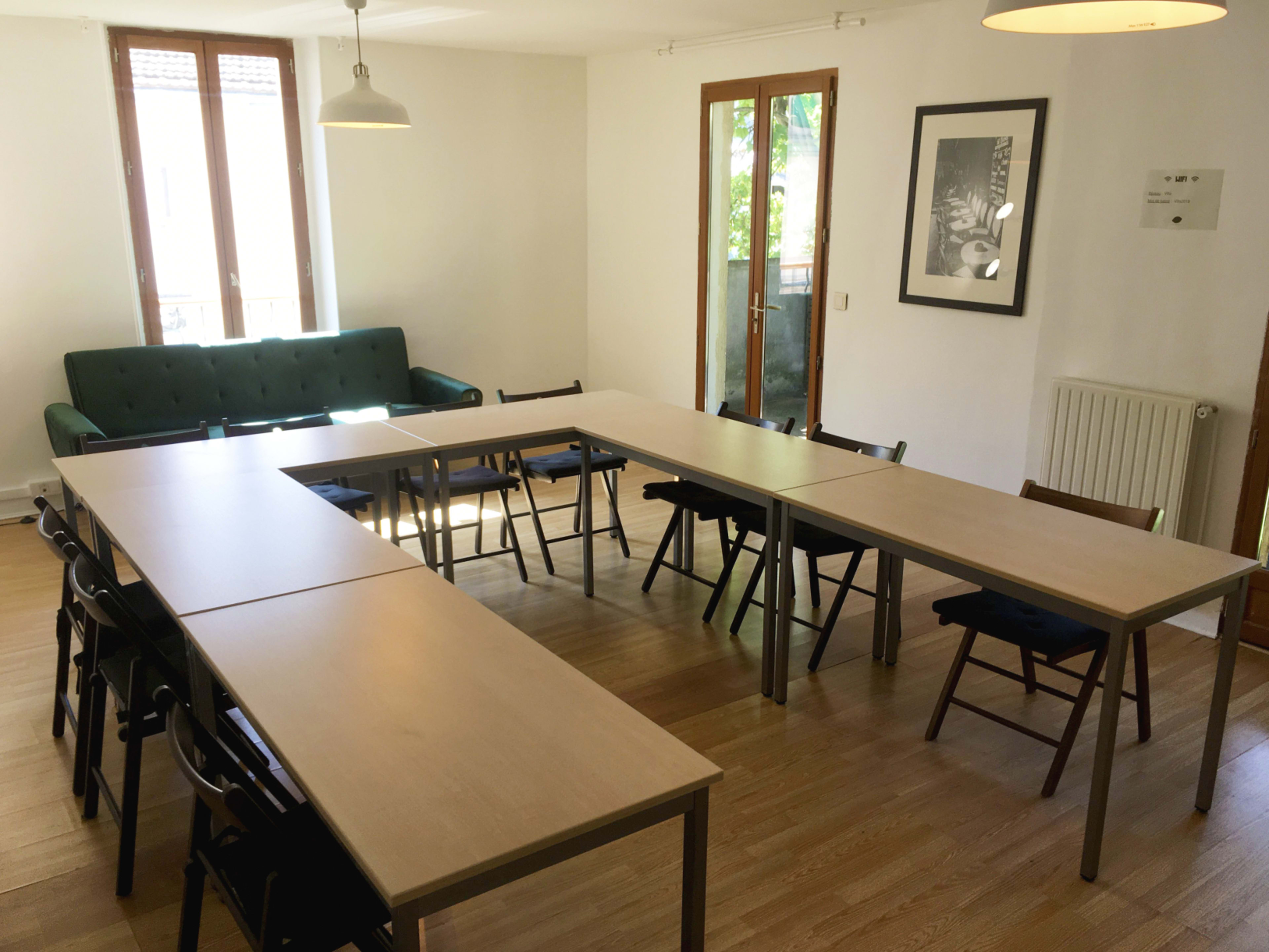 A room is set up with several tables arranged in a U-shape, surrounded by chairs and featuring windows that allow natural light.