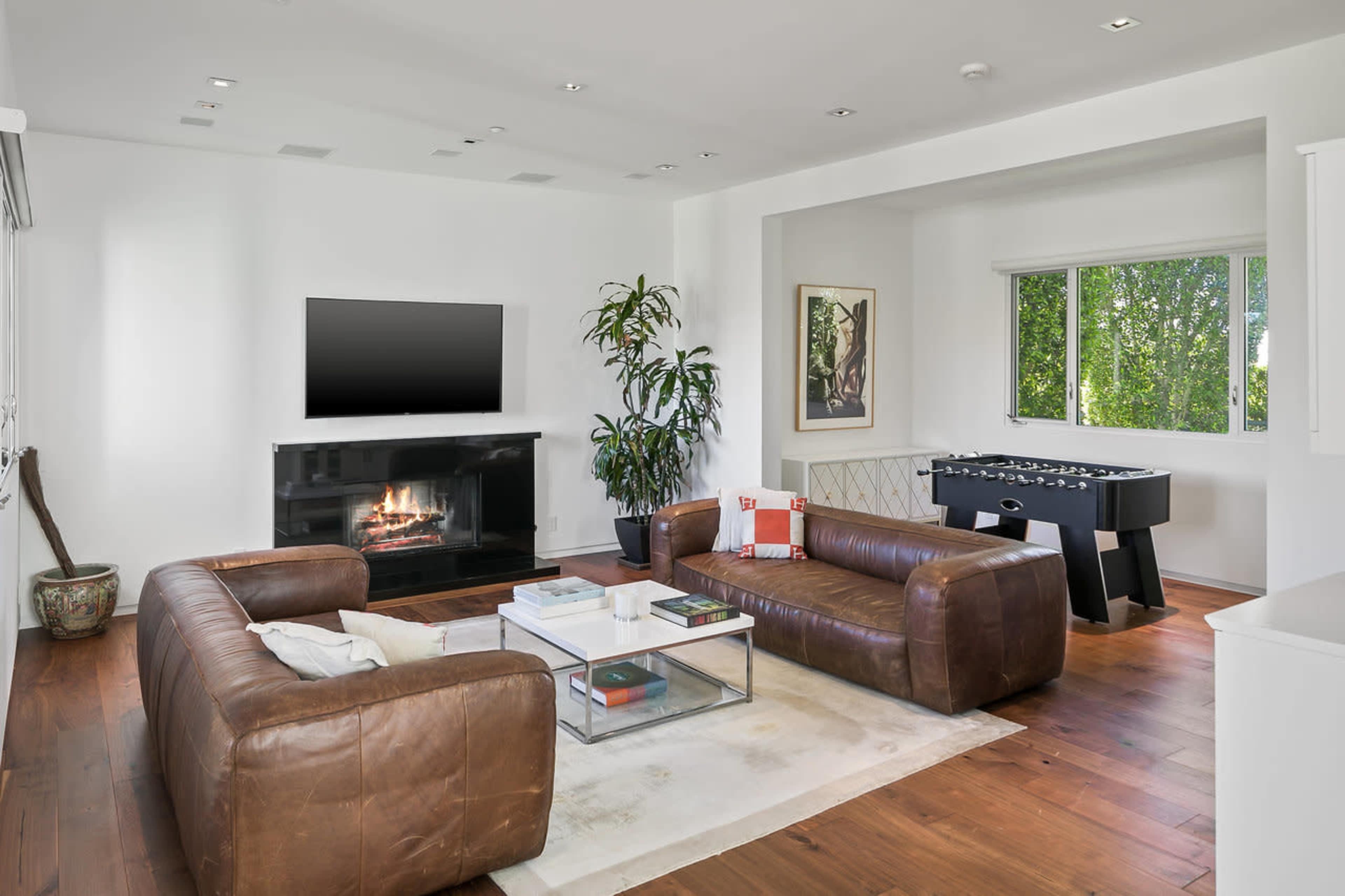 A bright living room features a leather sofa, a coffee table, a modern fireplace, and a foosball table near a window.