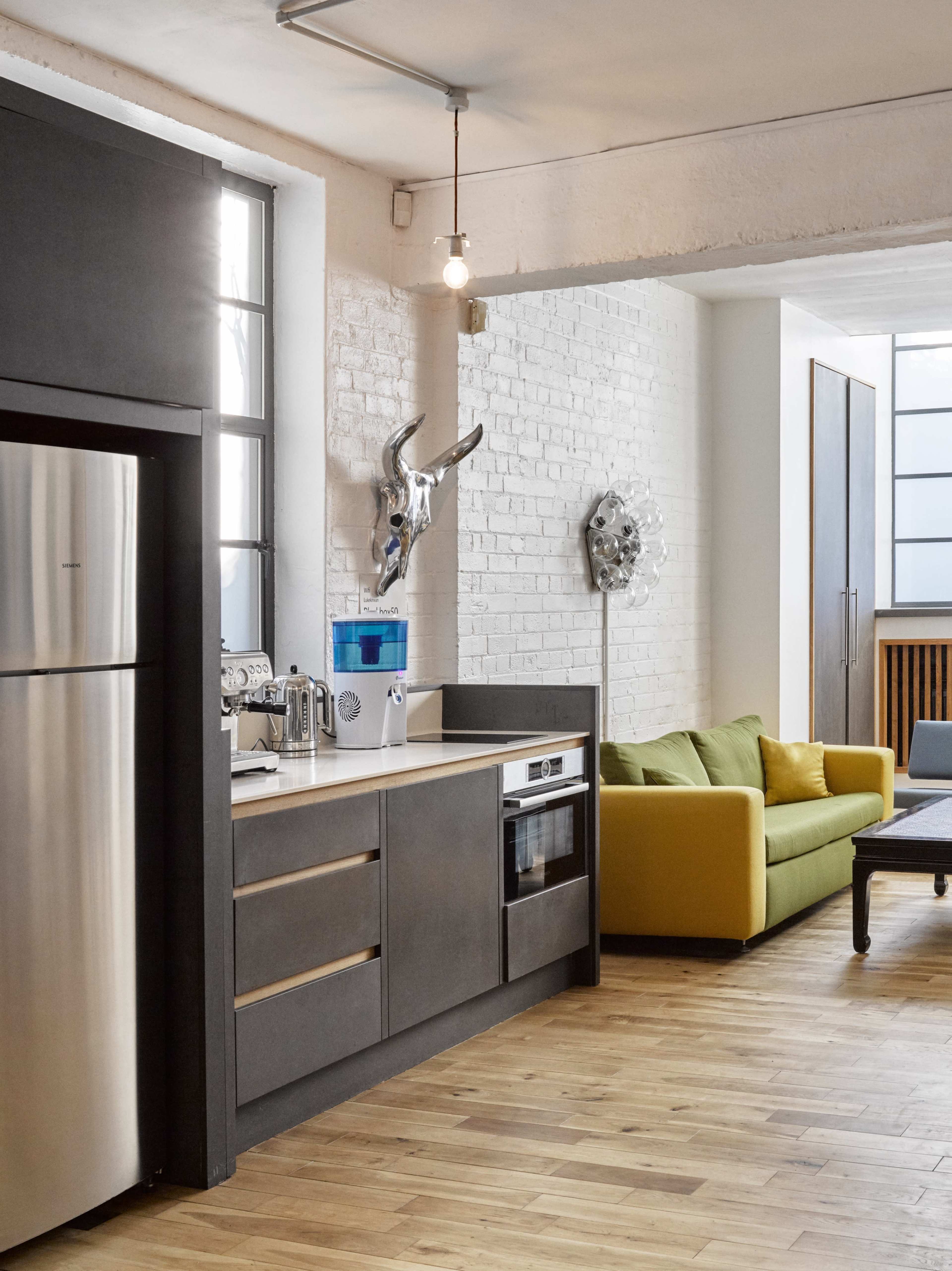 The image depicts a modern kitchen with stainless steel appliances and dark cabinetry, adjacent to a bright living area featuring a yellow sofa and artistic wall decor.