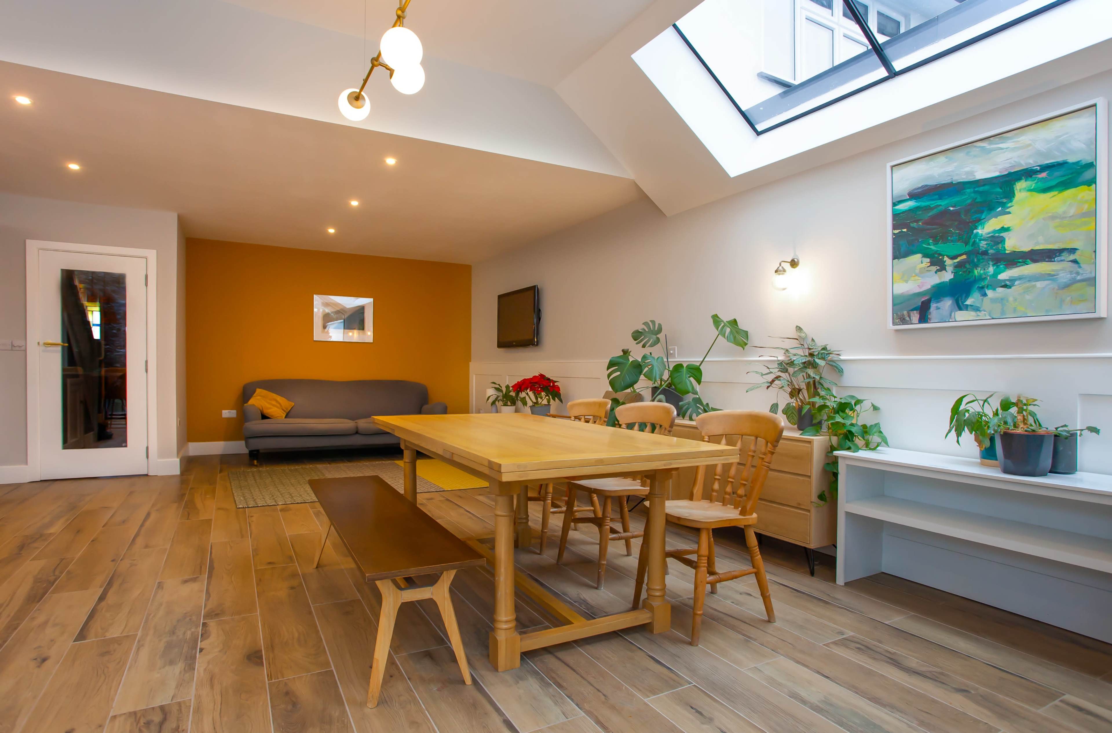 The image shows a modern dining area with a wooden table and chairs, a sofa against a wall, indoor plants, and a large skylight above.