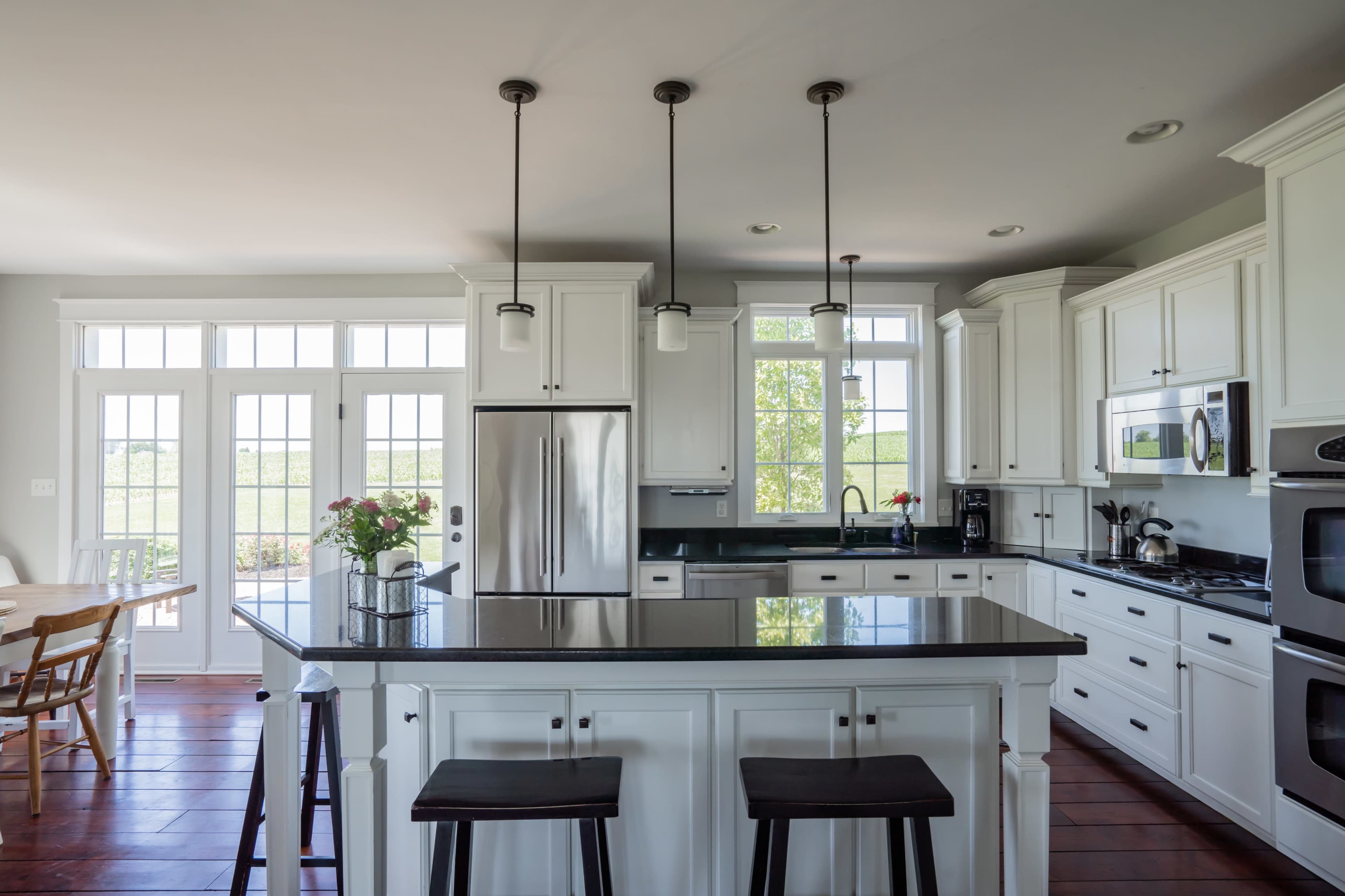 A bright kitchen features white cabinets, a dark countertop, and large windows overlooking a garden.