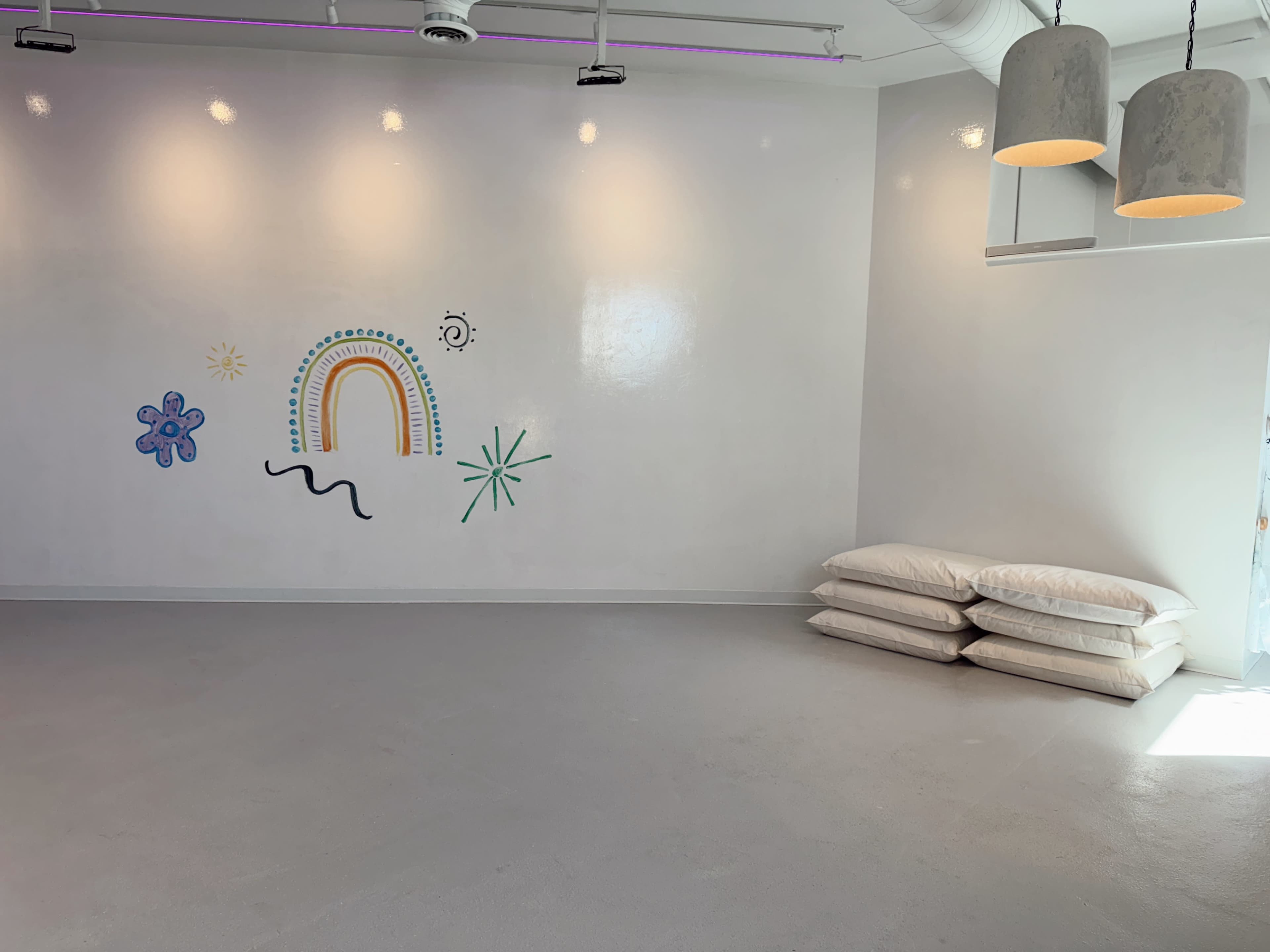 The image shows a spacious, empty room with a decorated wall featuring a rainbow and some abstract designs, along with a stack of pillows in one corner.