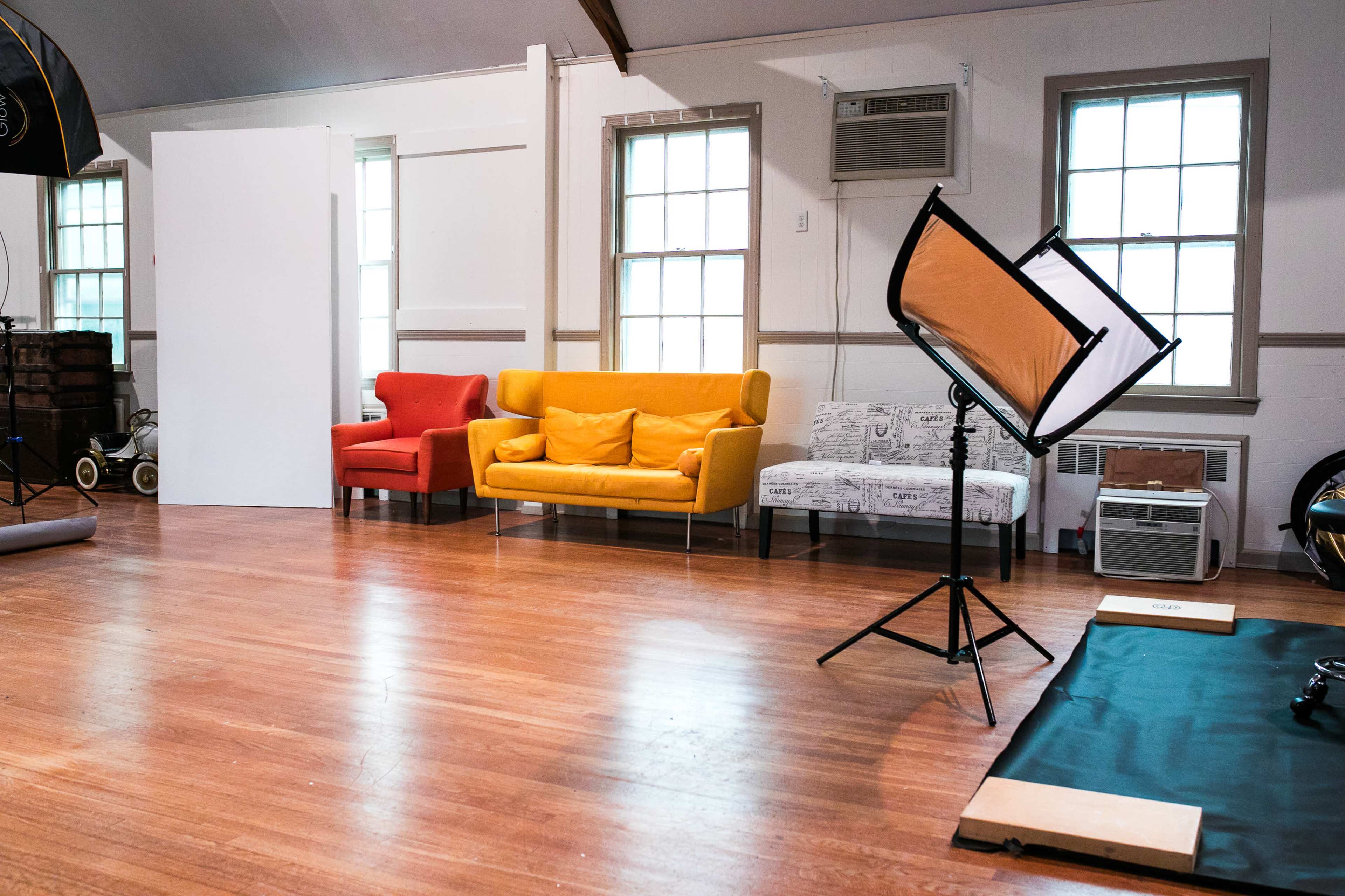 A spacious studio with wooden flooring, featuring two colorful sofas, a lighting stand, and large windows.
