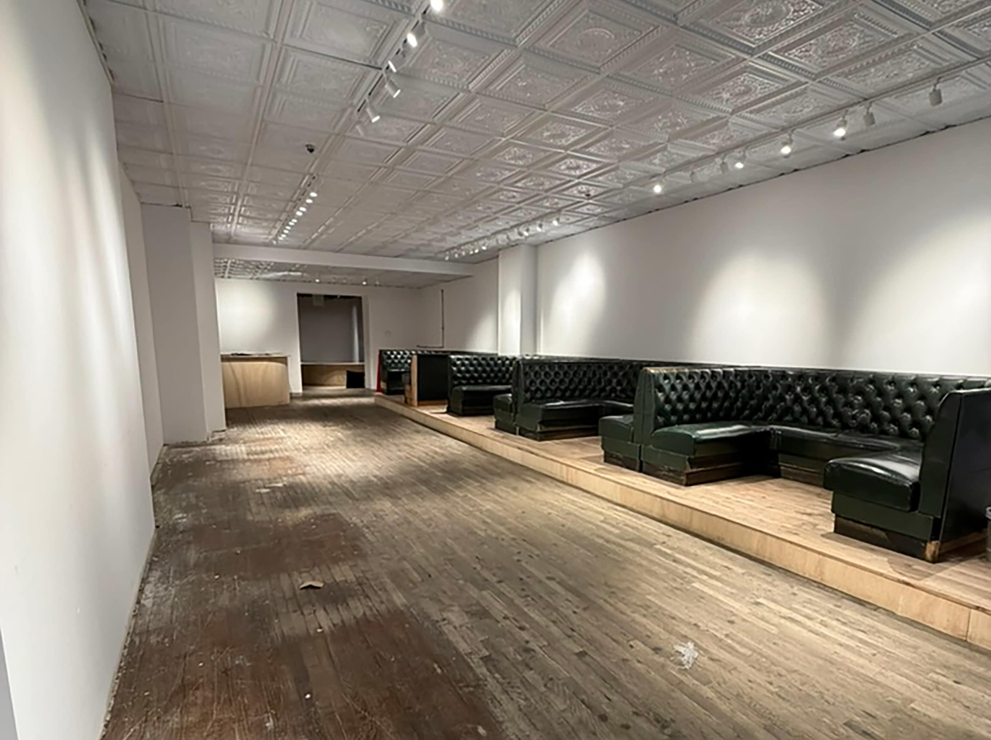 The image shows a spacious, empty room with wooden flooring, featuring dark green tufted seating along the walls and recessed lighting illuminating the area.