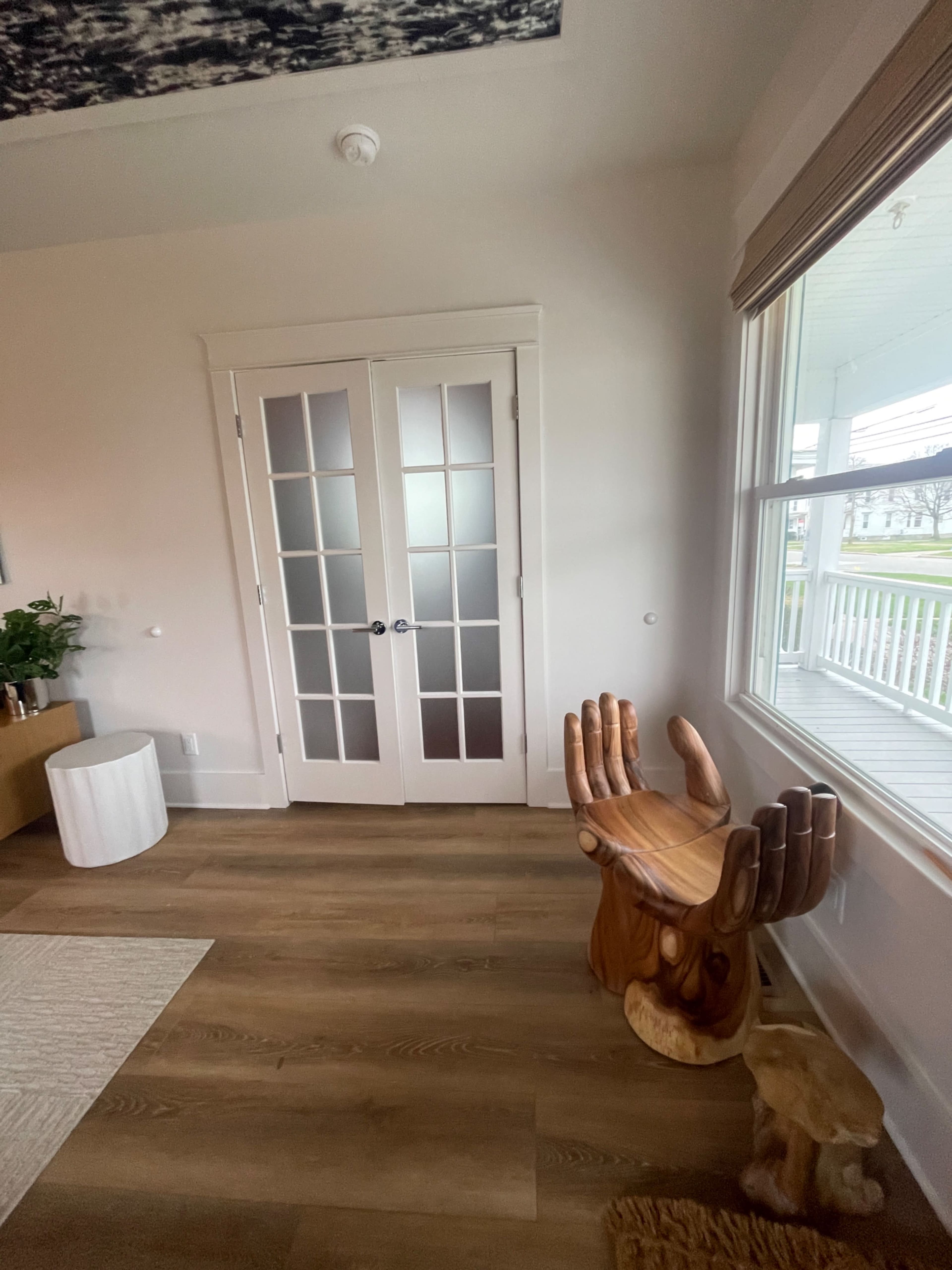 A corner of a room featuring a wooden hand-shaped chair next to a pair of glass-panel double doors and a window.