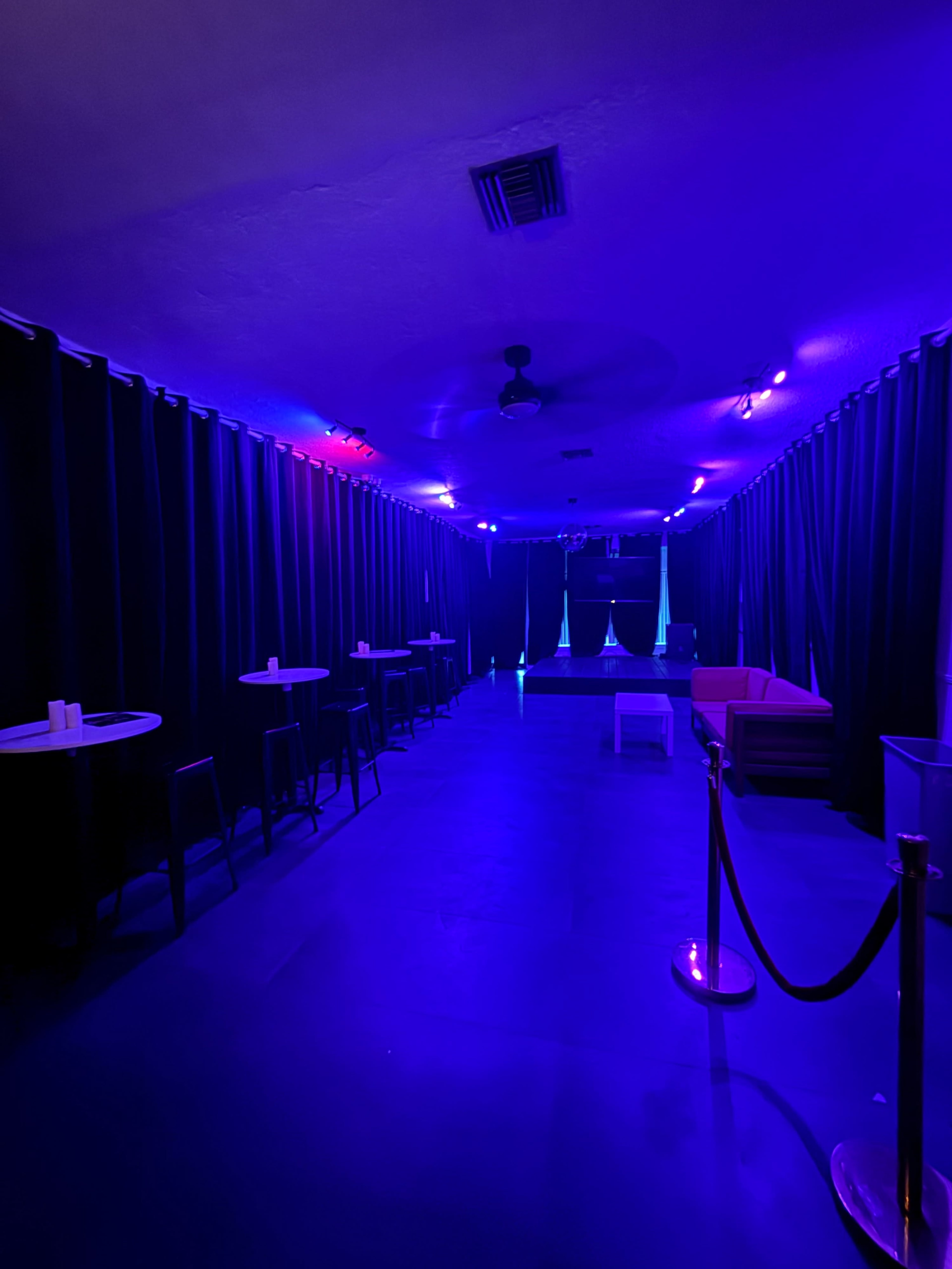 The image shows a dimly lit room featuring purple lighting, black curtains along the walls, and scattered tables and chairs.