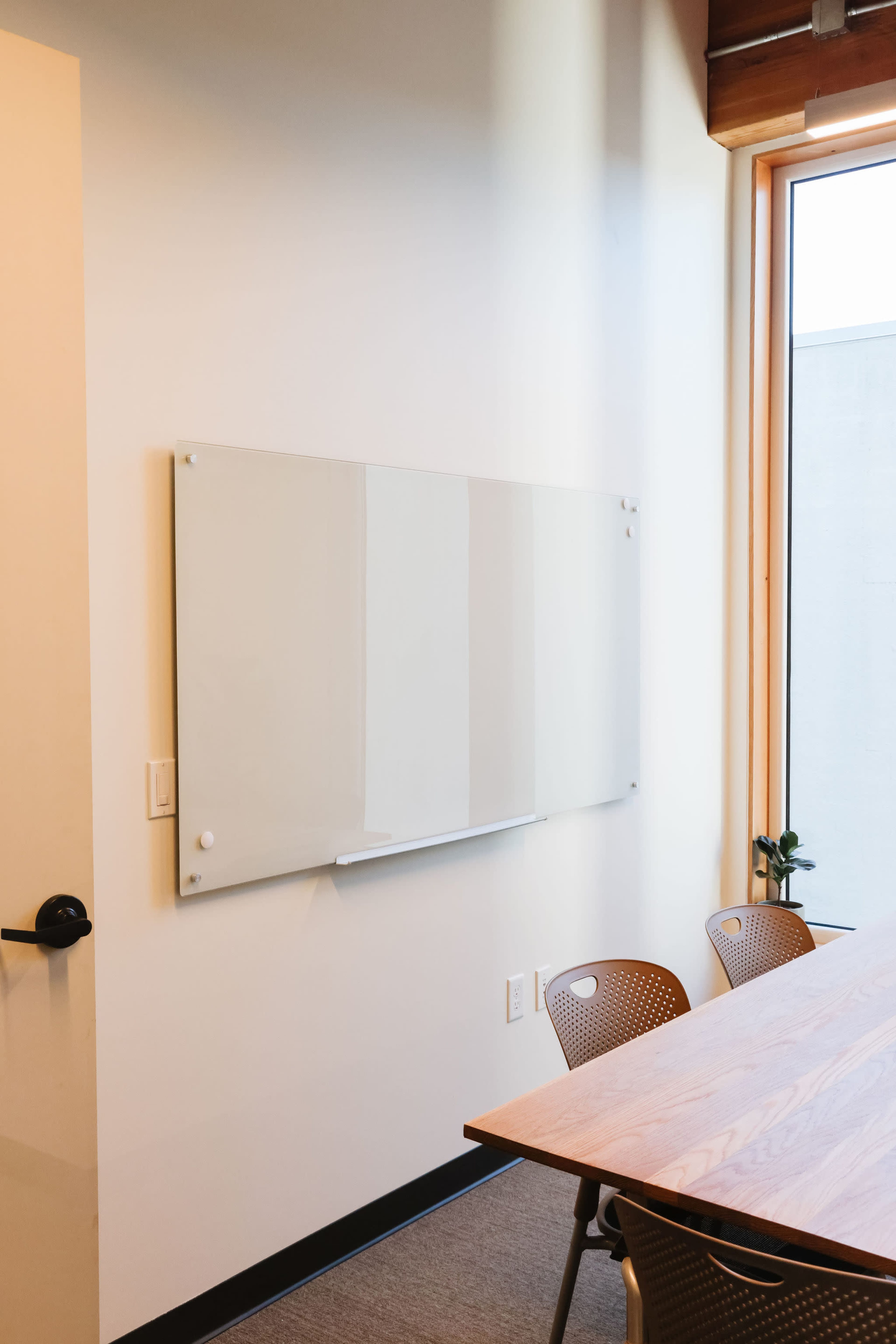A blank whiteboard is mounted on a light wall next to a large window, overlooking a table with several chairs.