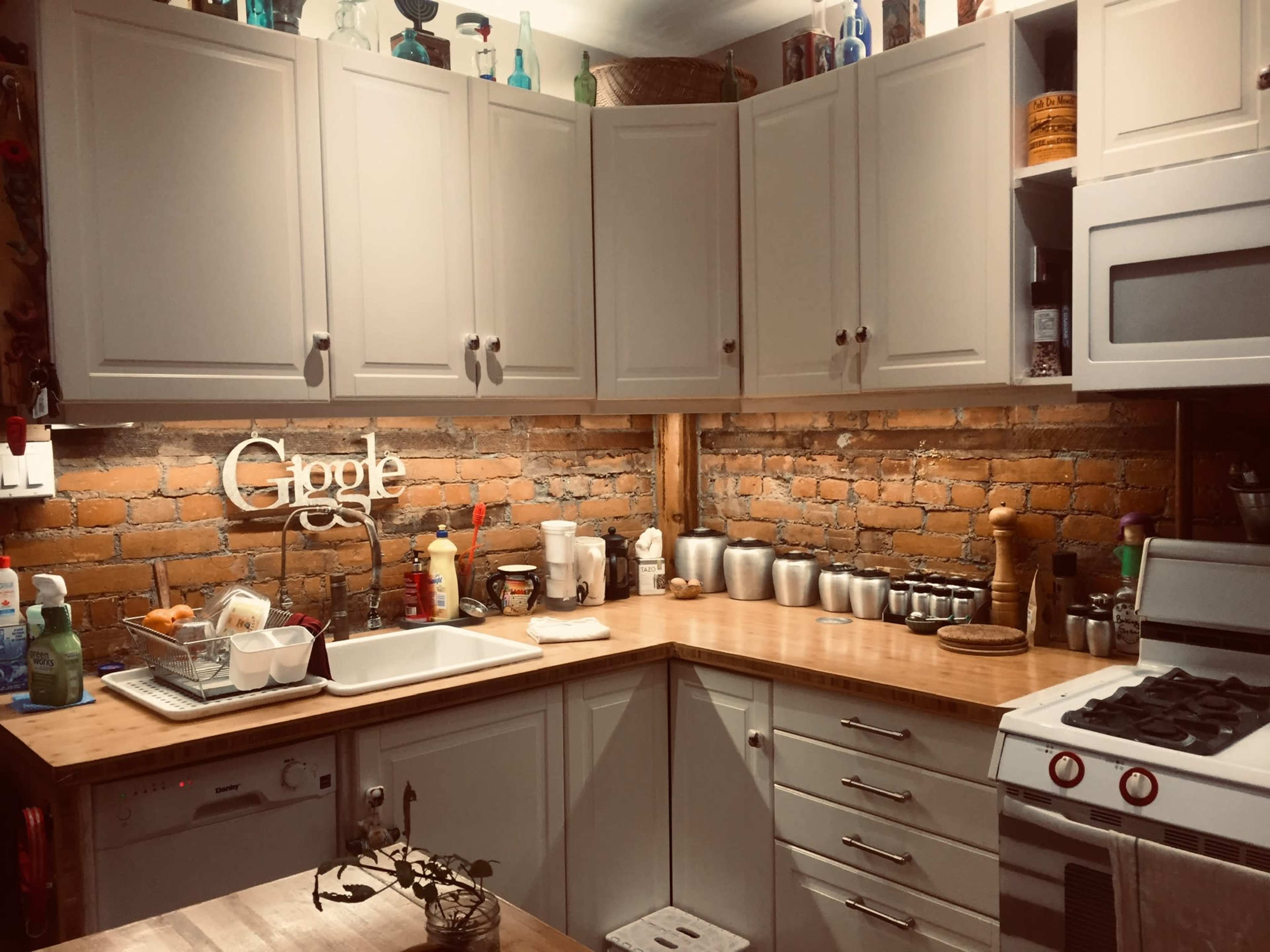 A kitchen features wooden countertops, white cabinetry, and exposed brick walls, with various utensils and appliances arranged on the surfaces.