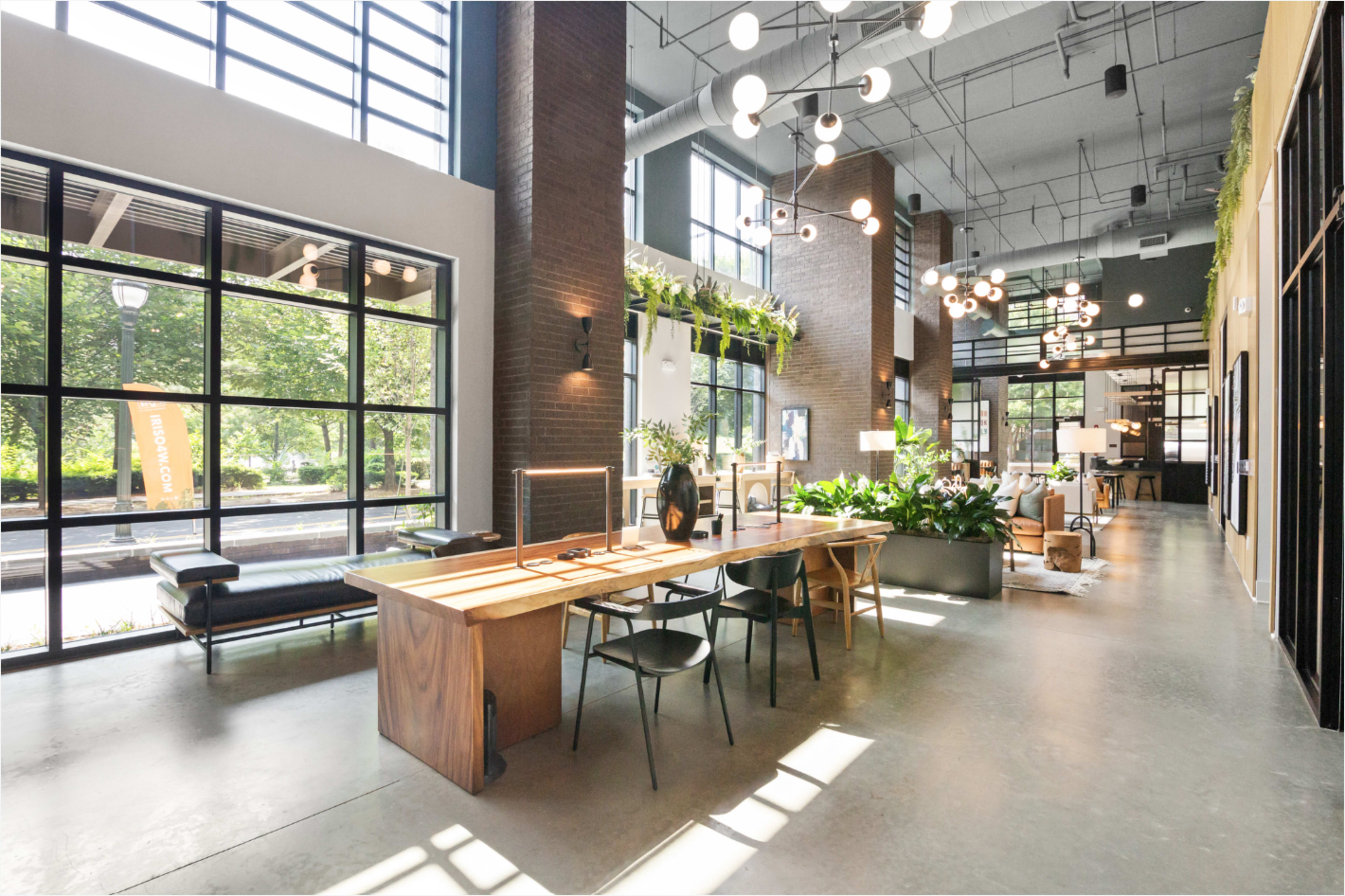 The image shows a spacious, modern interior with large windows, a long wooden table, dark chairs, and various indoor plants.