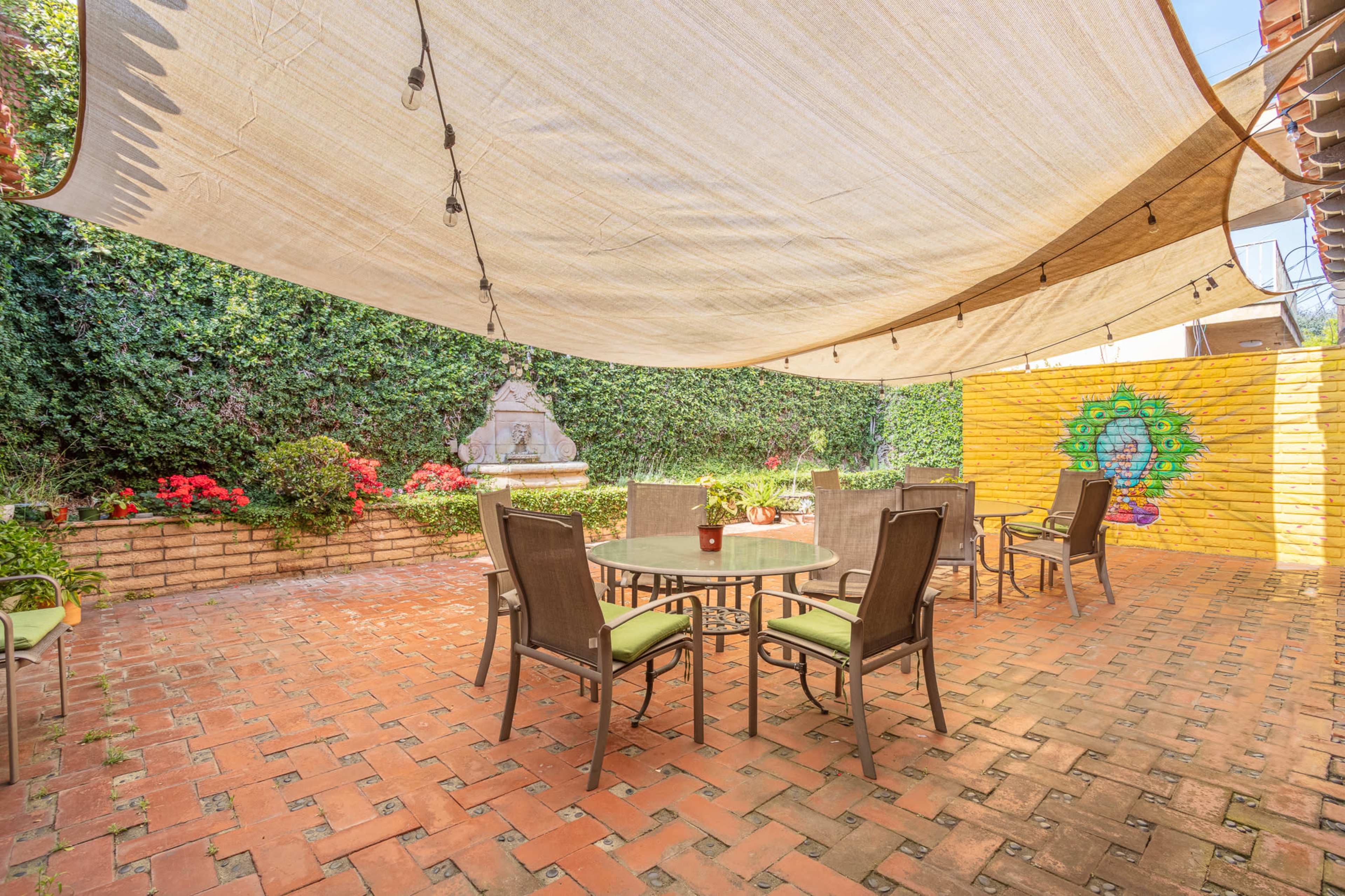 A patio area is shaded by a large canopy and features a round table with six chairs, surrounded by vibrant flowers and a mural on the wall.