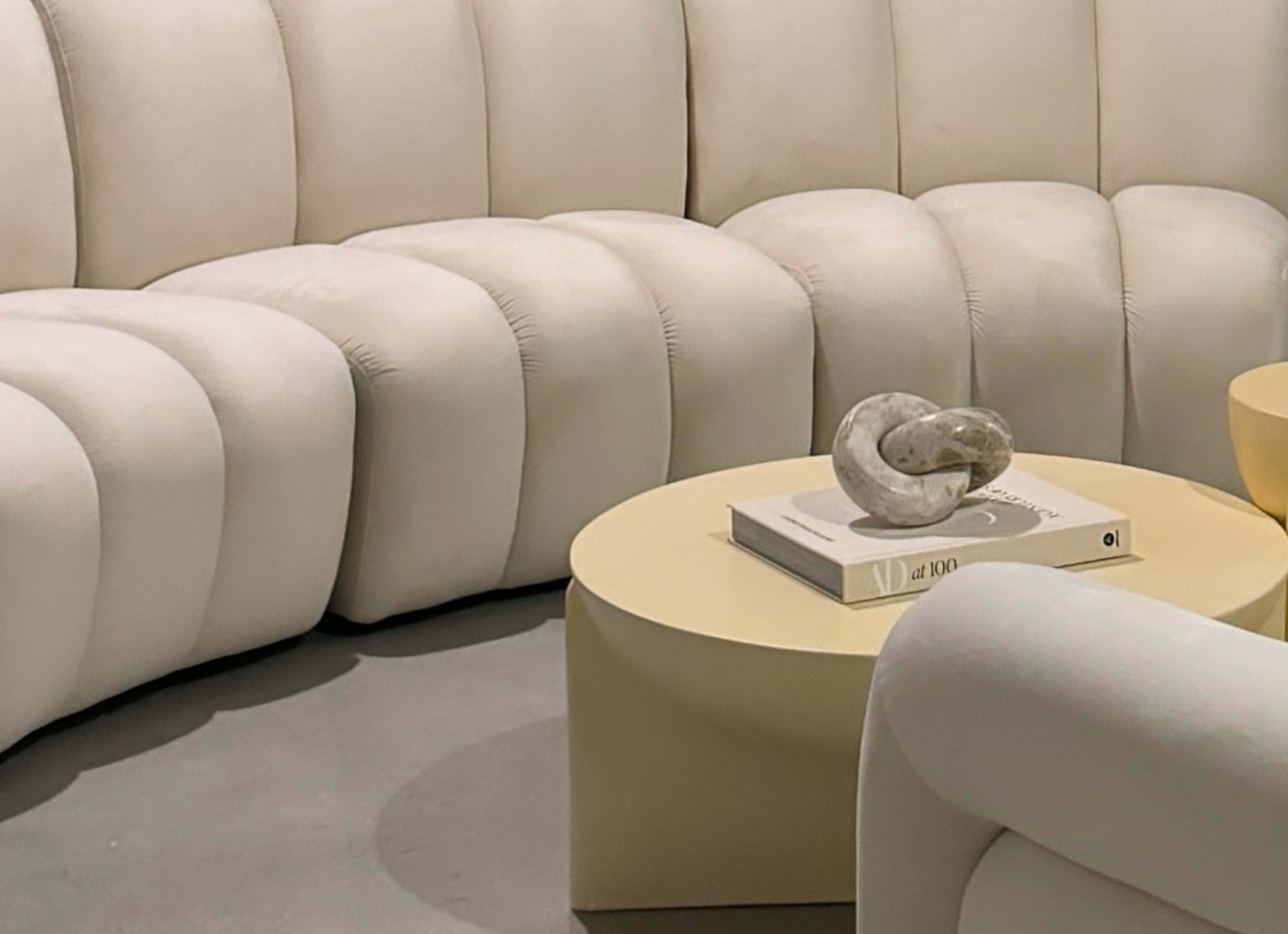 The image shows a curved, cream-colored sectional sofa alongside a round, light-colored coffee table with a decorative object and a stacked magazine.