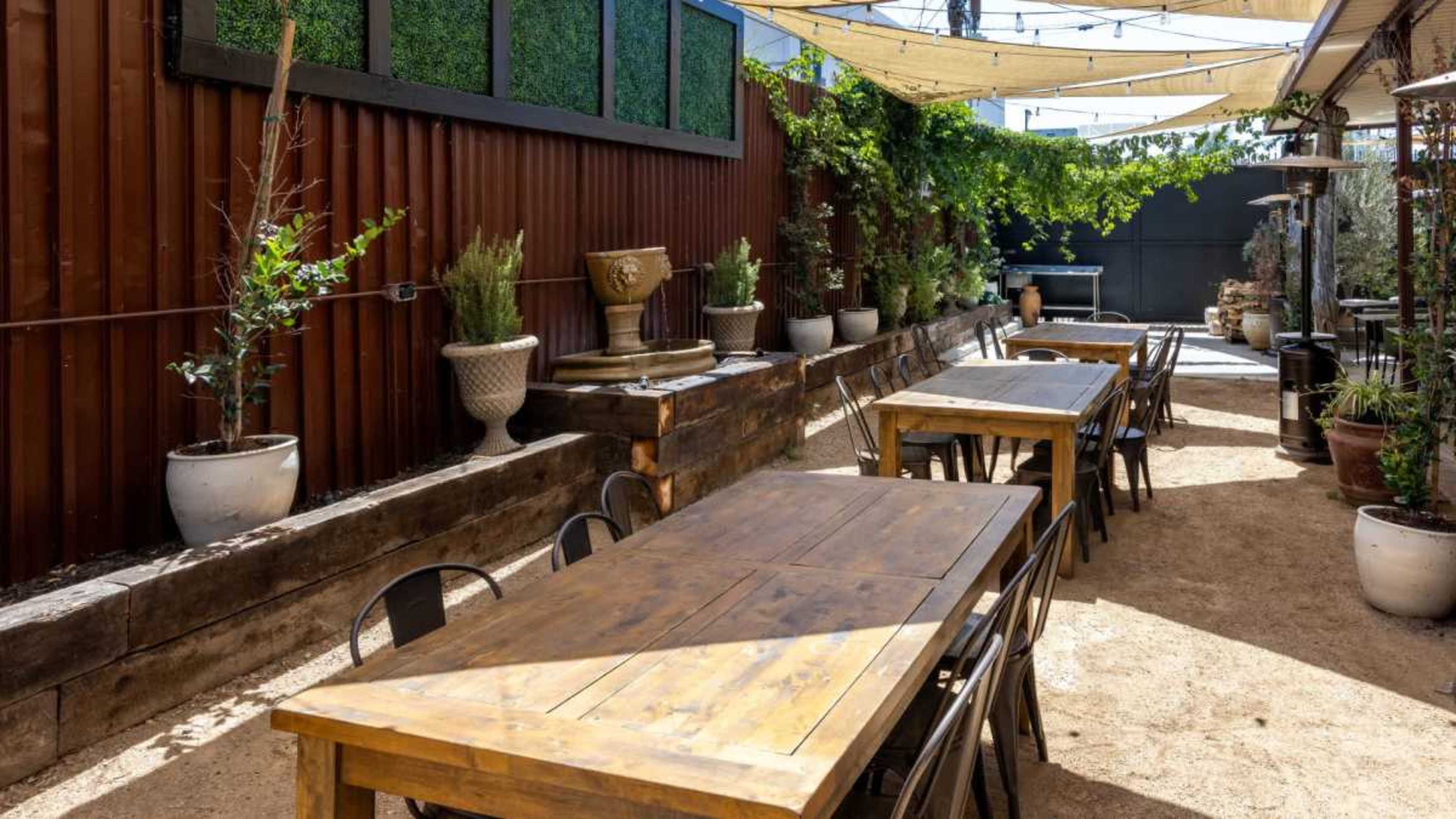 The image shows an outdoor dining area with wooden tables arranged along a gravel path, surrounded by potted plants and a decorative fountain.