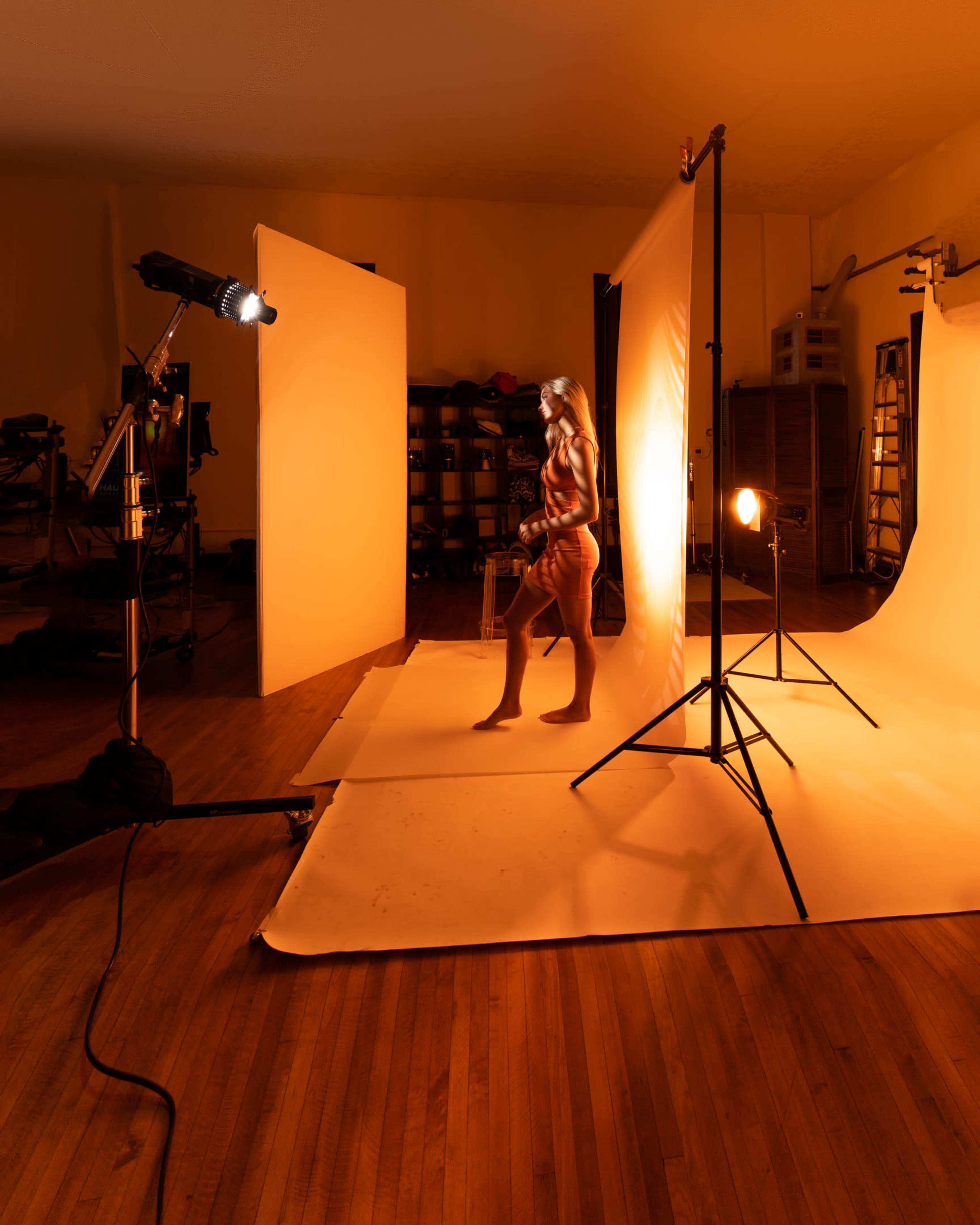 A model stands on a photo shoot set with lighting equipment and backdrops arranged for a studio session.