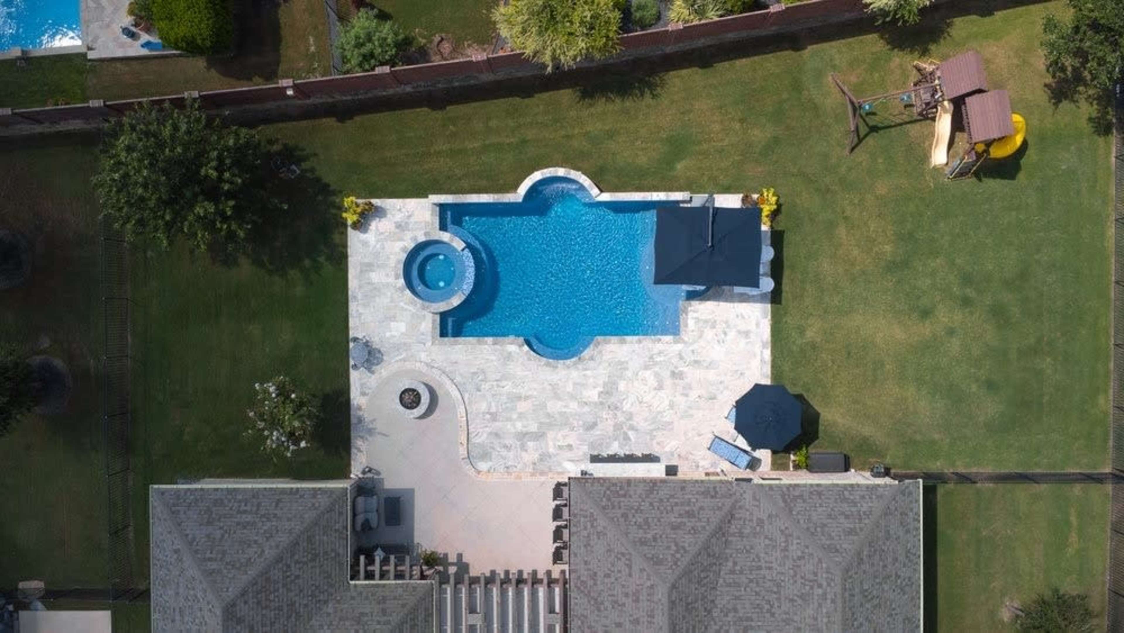 The image shows a backyard with a swimming pool featuring a hot tub, surrounded by a stone patio and lawn, with a playset in the corner.