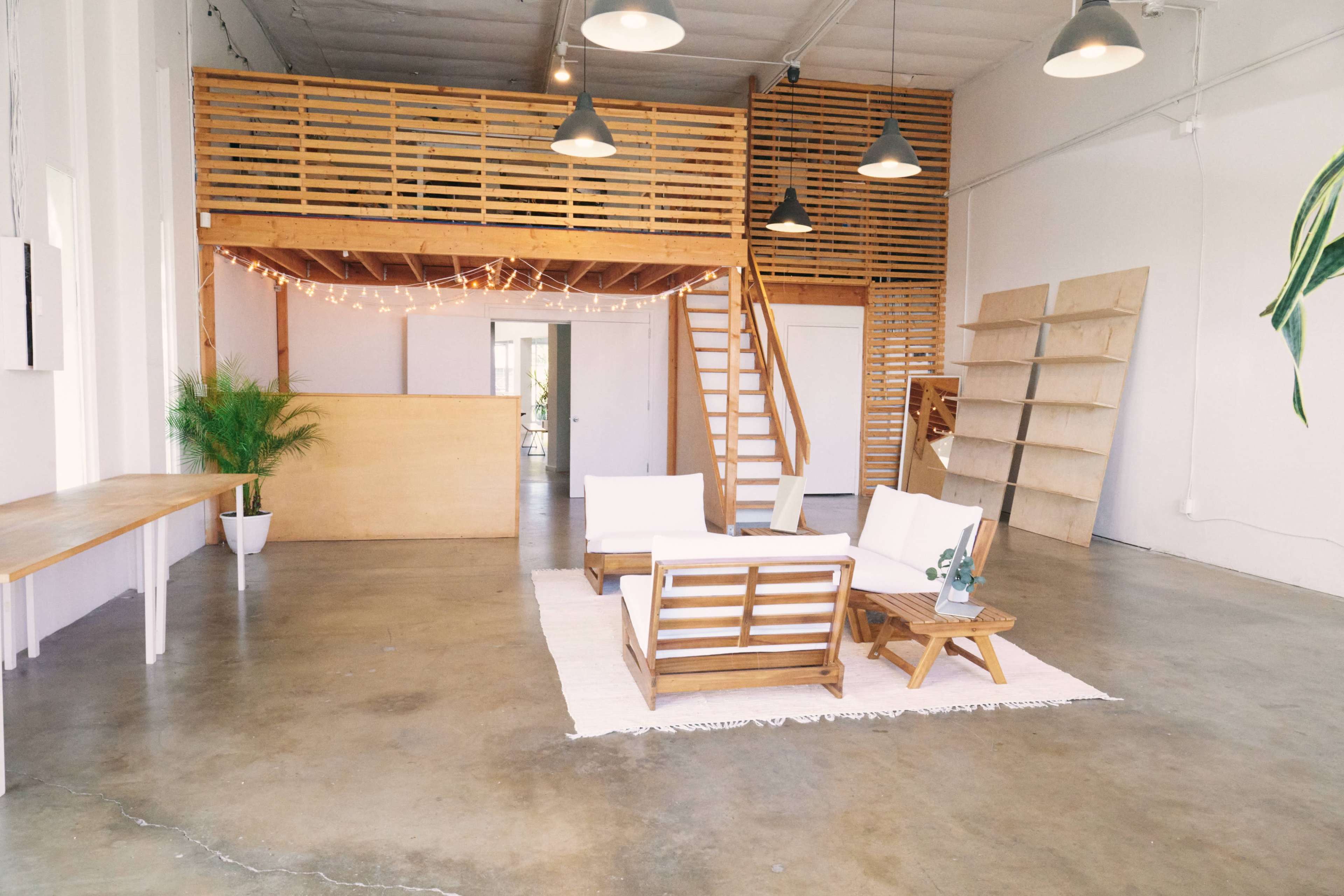 A spacious interior with wooden accents, featuring a seating area with white couches, a staircase leading to a mezzanine, and open shelving along the wall.
